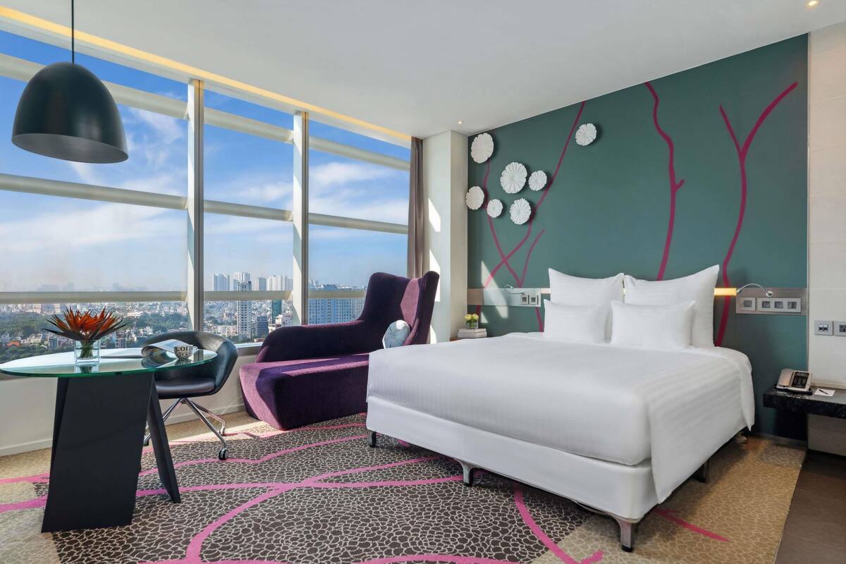 Ein modernes Hotelzimmer mit einem großen Bett, einem Schreibtisch mit Stuhl, Blick auf die Stadt durch große Fenster und einer Wand mit abstrakten Mustern.