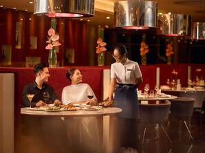 Interior de un restaurante con mesas redondas, asientos tapizados en rojo y personal de servicio atendiendo a los clientes.