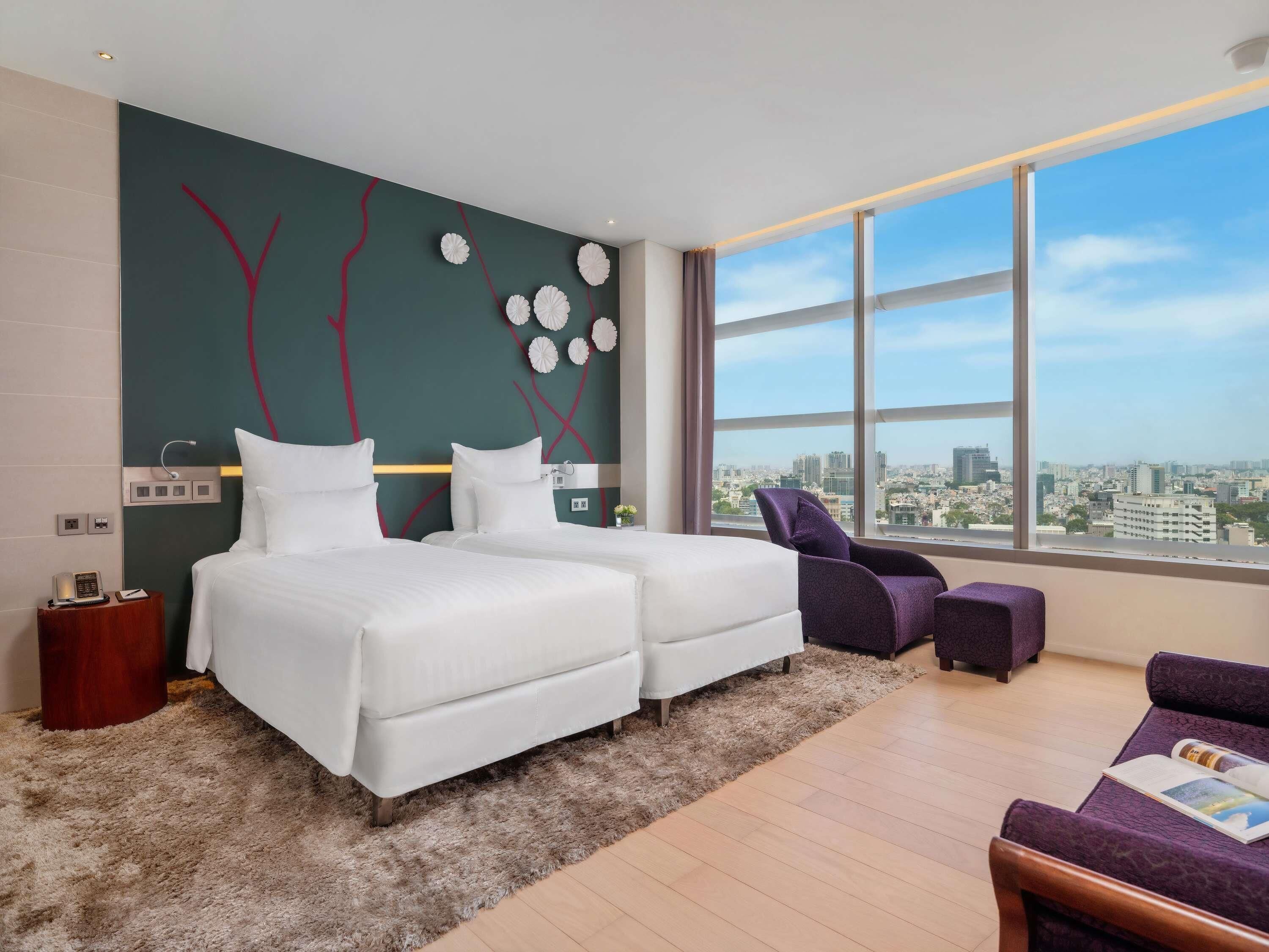 A modern hotel room with a large window offering a city view, a bed with white linens, a purple armchair, and a small side table with a phone and a book.