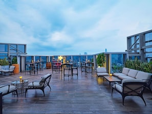 A rooftop bar with tables and chairs, a bar counter, and a city view.