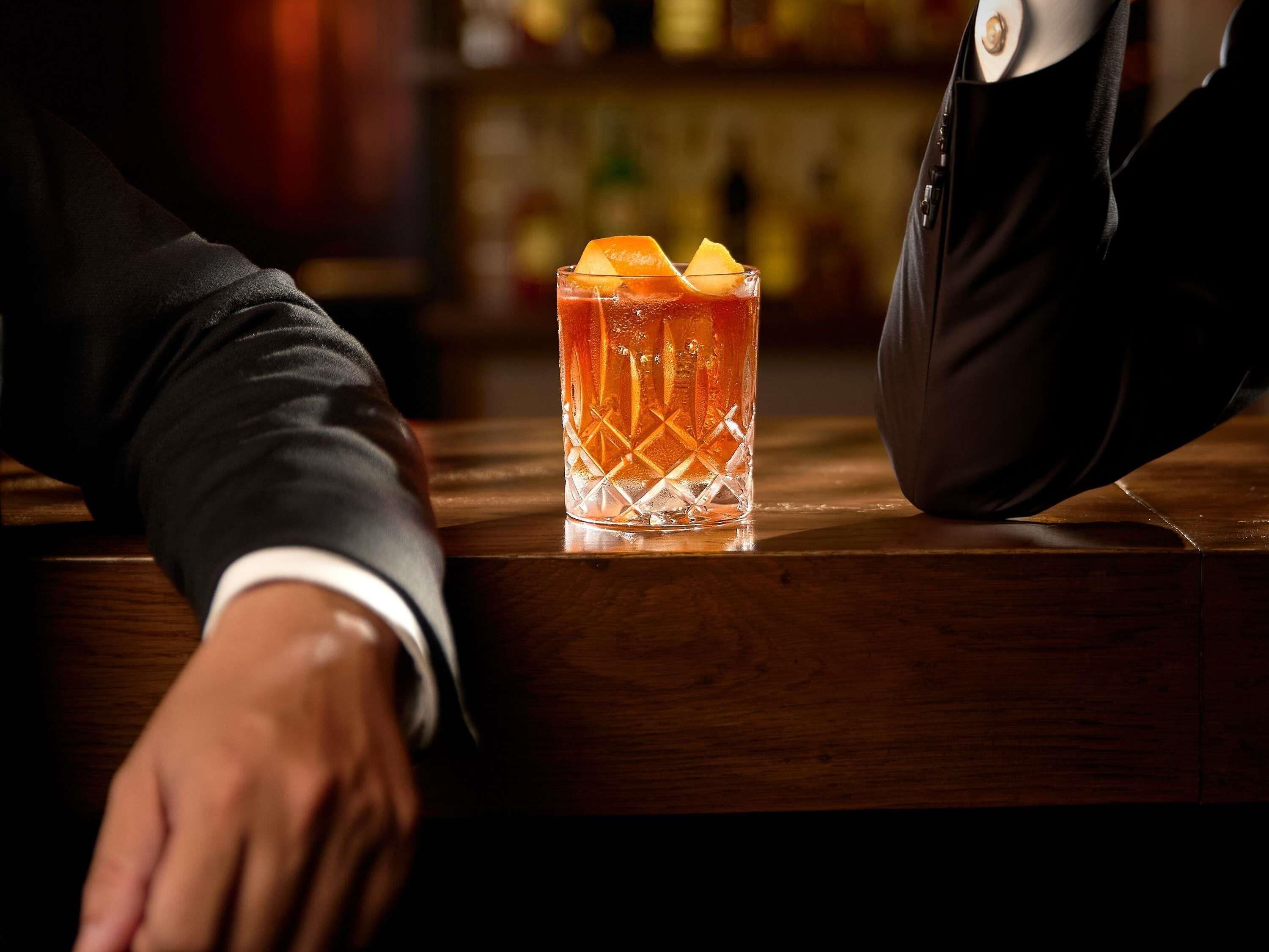 Eine Bar mit einem Glas bernsteinfarbenen Getränks, verziert mit Zitrusfrüchten, einer Holzbar und einer Hand, die darauf ruht.