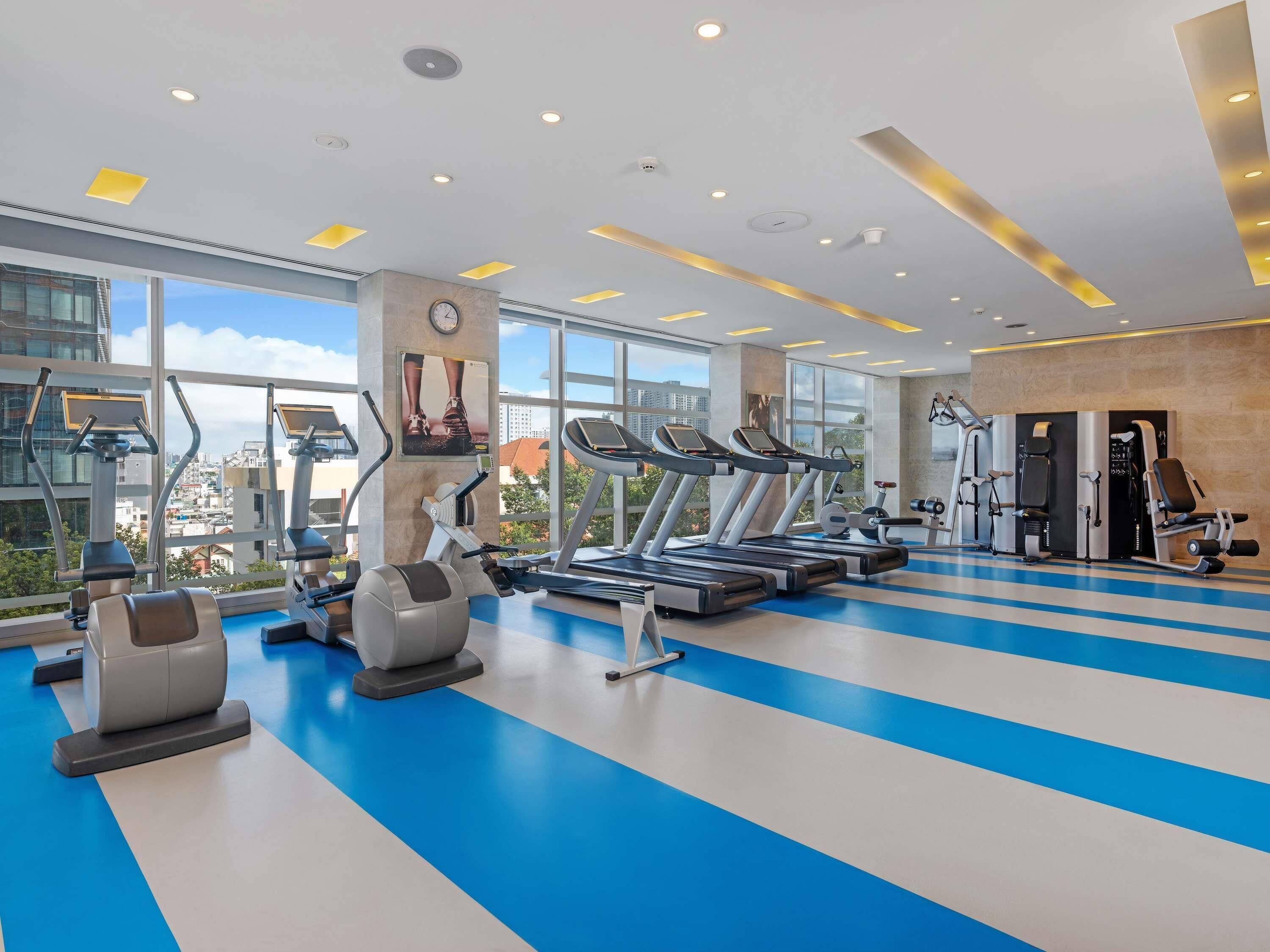 A modern gym with treadmills, ellipticals, and weight machines, large windows offering city views, and a blue and white floor pattern.