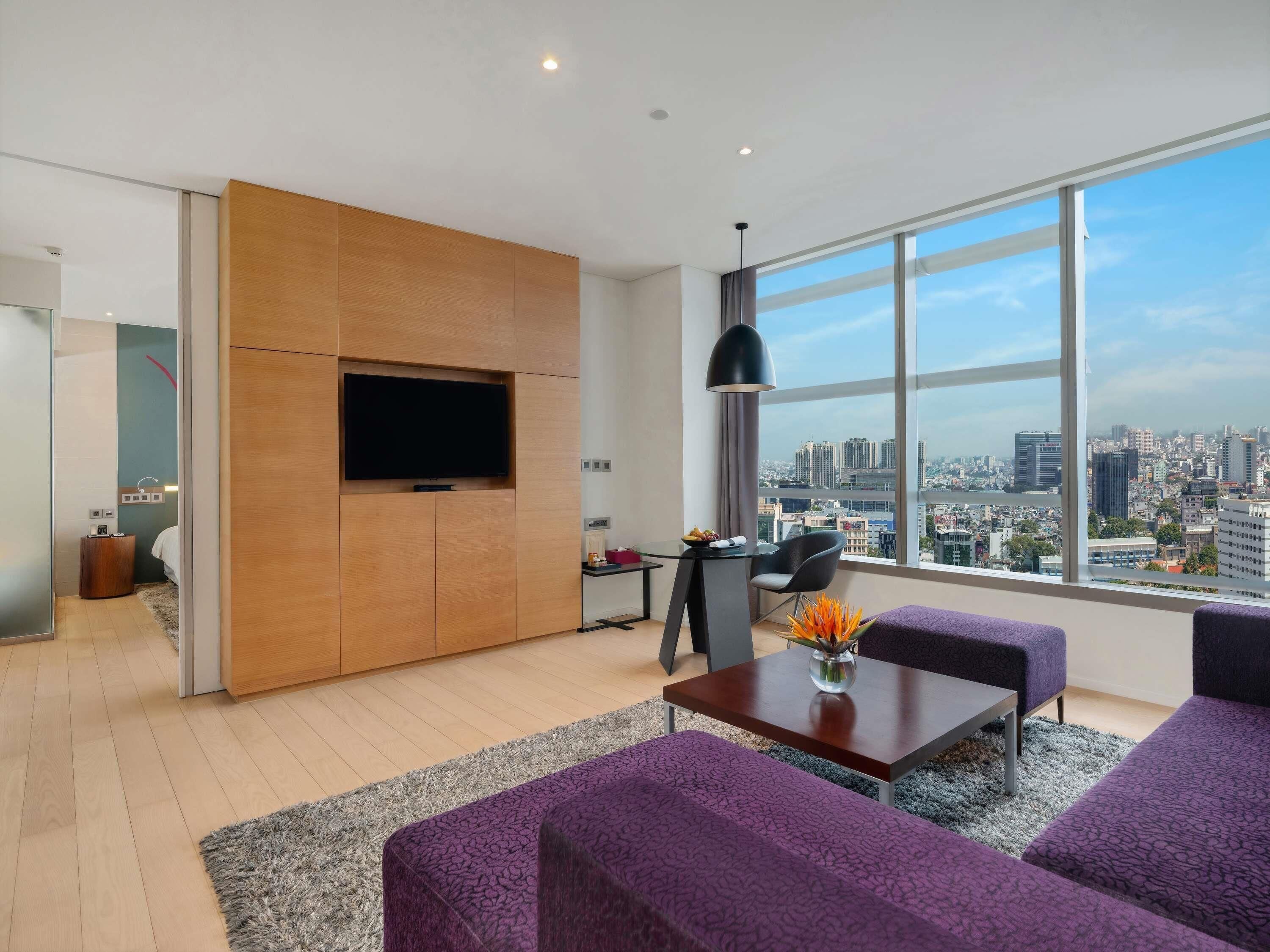 A modern living room with a large TV, purple sofas, a wooden cabinet, and a city view through large windows.