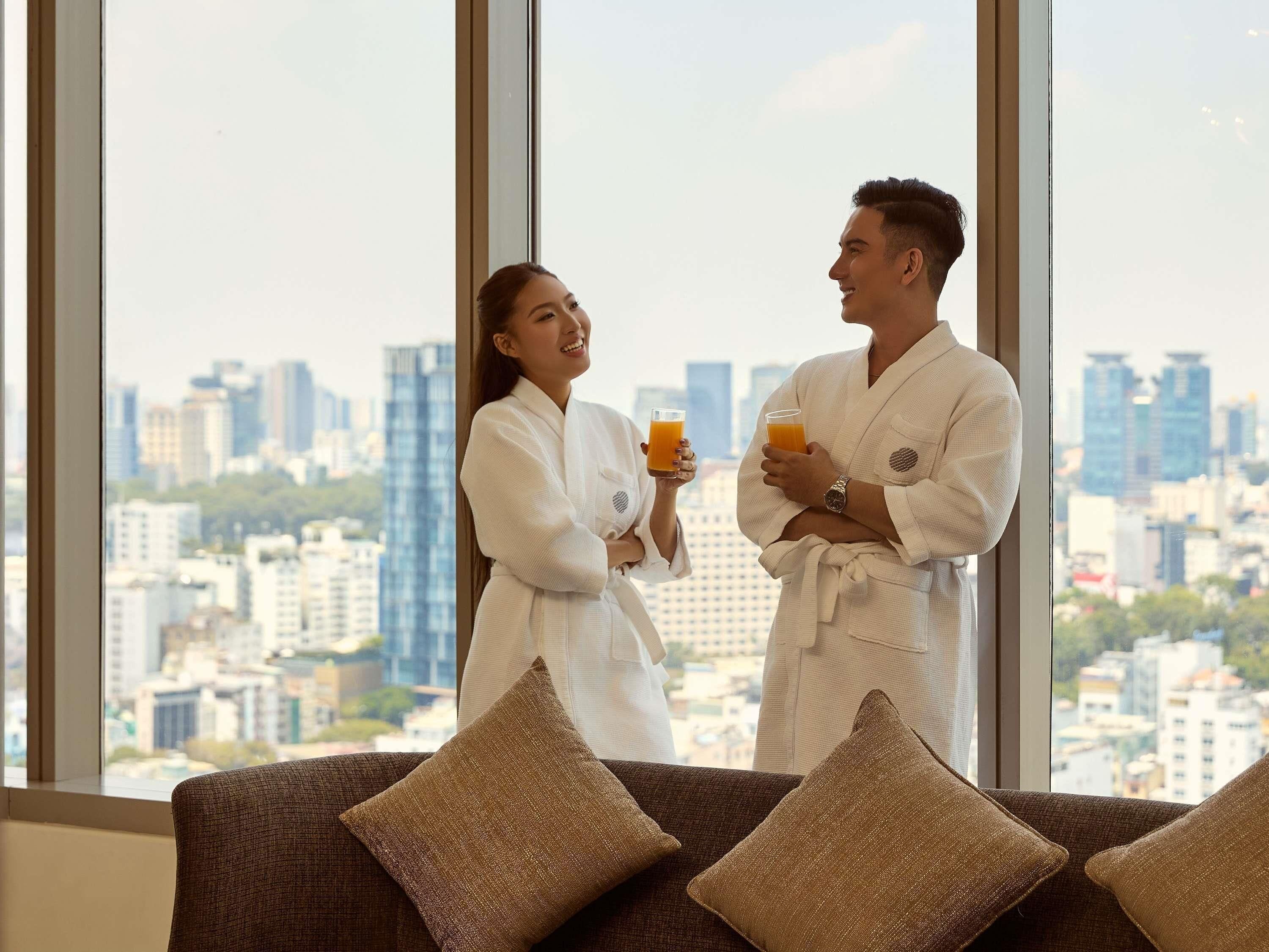 Two people in white bathrobes holding orange beverages, standing by a window with a cityscape view.