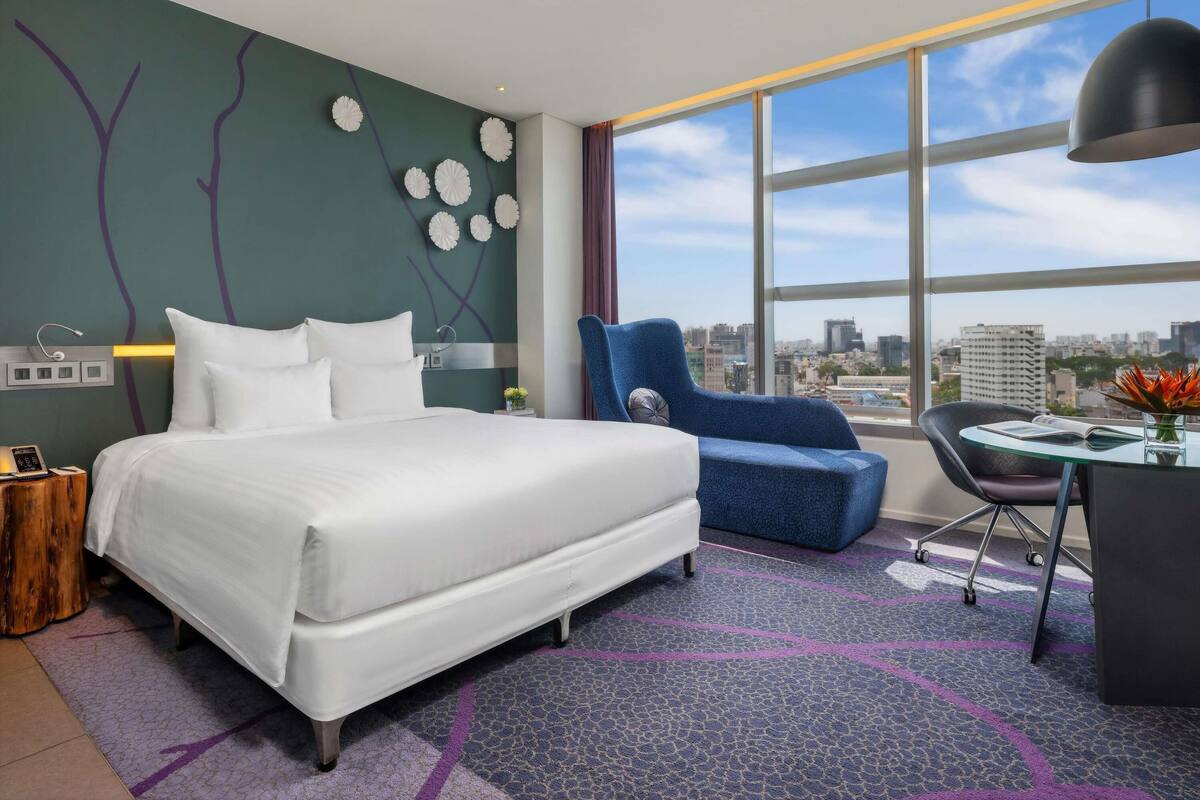 Ein modernes Hotelzimmer mit einem großen Bett, einem blauen Sessel, einem Schreibtisch mit Lampe und Blick auf die Stadt.