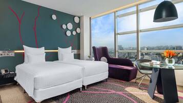 A modern hotel room with a large bed, a purple armchair, a glass table with a vase, and a city view through large windows.