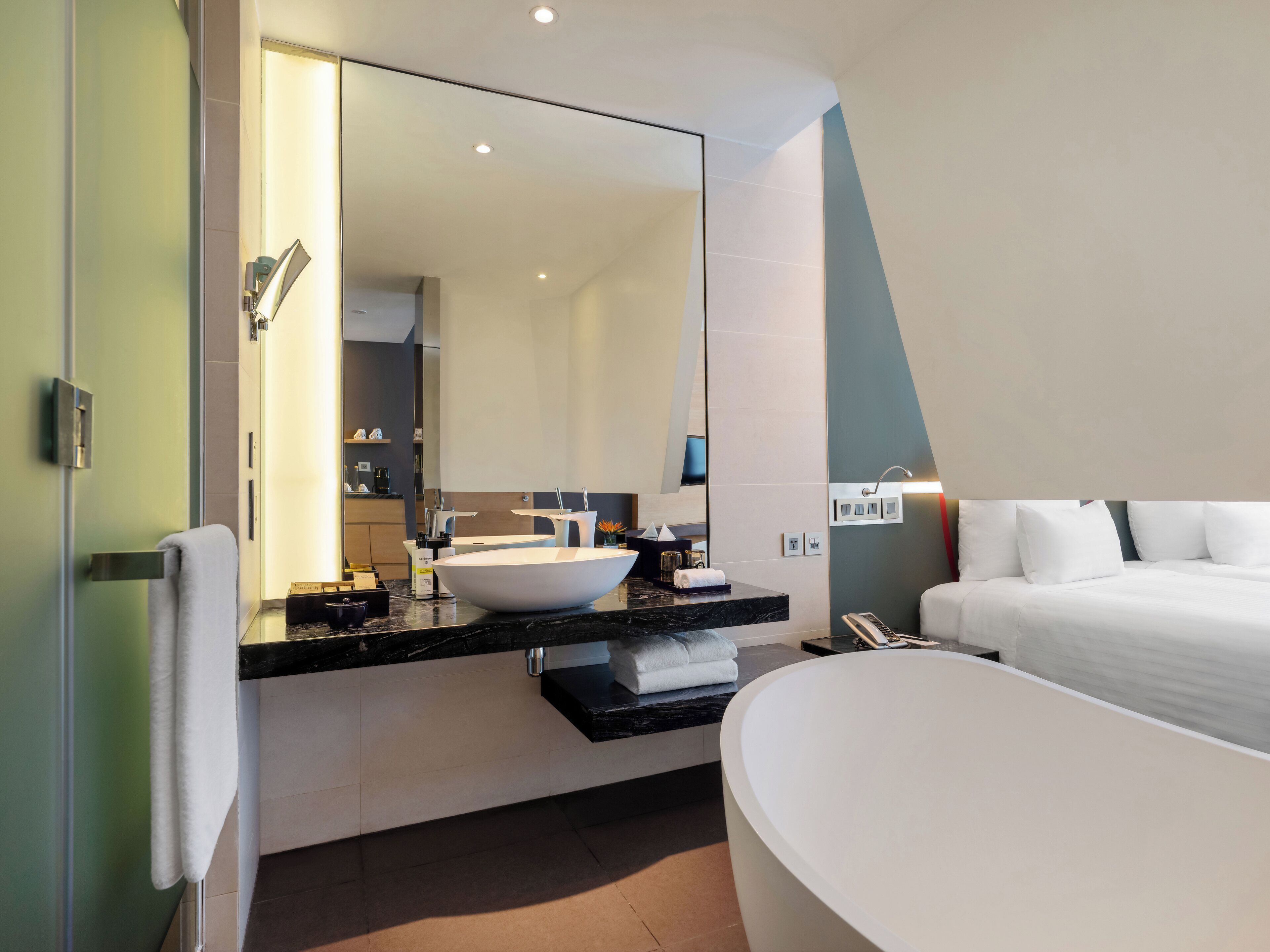 Ein modernes Badezimmer mit großem Spiegel, freistehender Badewanne, Waschtisch und einem ordentlich bezogenen Bett.