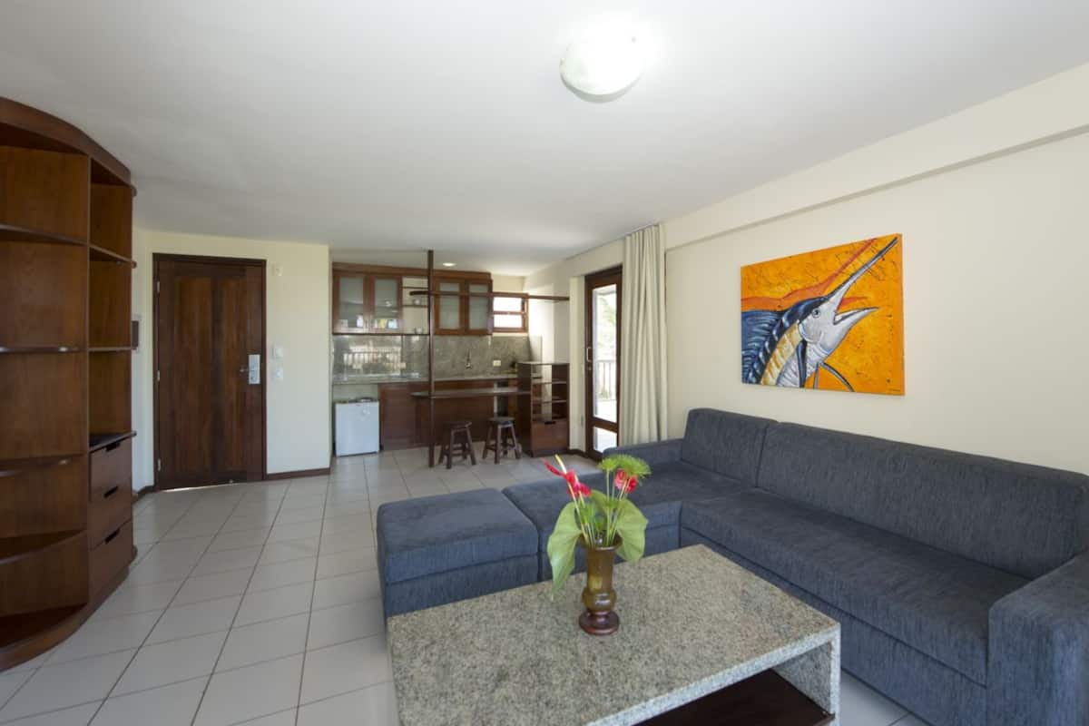 A living room with a grey sofa, a coffee table with a vase of flowers, and a kitchen in the background.