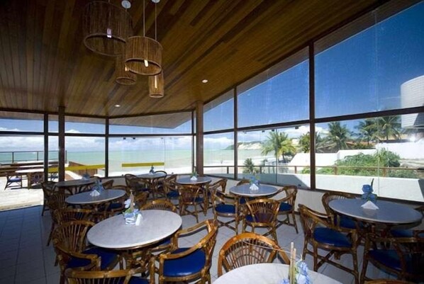 A restaurant with round tables and wicker chairs, large windows offering a view of the beach and ocean. - Blue Marlin Apartments (Natal)