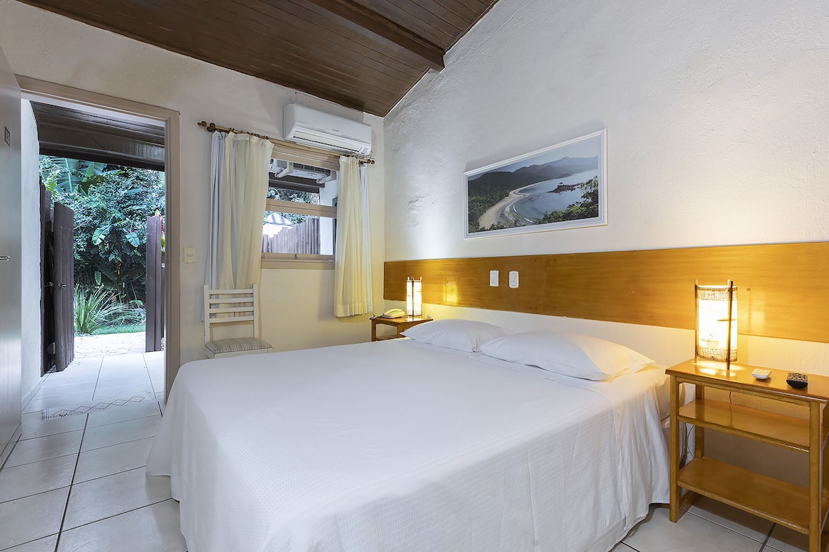 Une chambre d’hôtel avec un grand lit, une tête de lit en bois, une table de chevet avec une lampe, une petite table et une vue sur l’extérieur par une fenêtre.