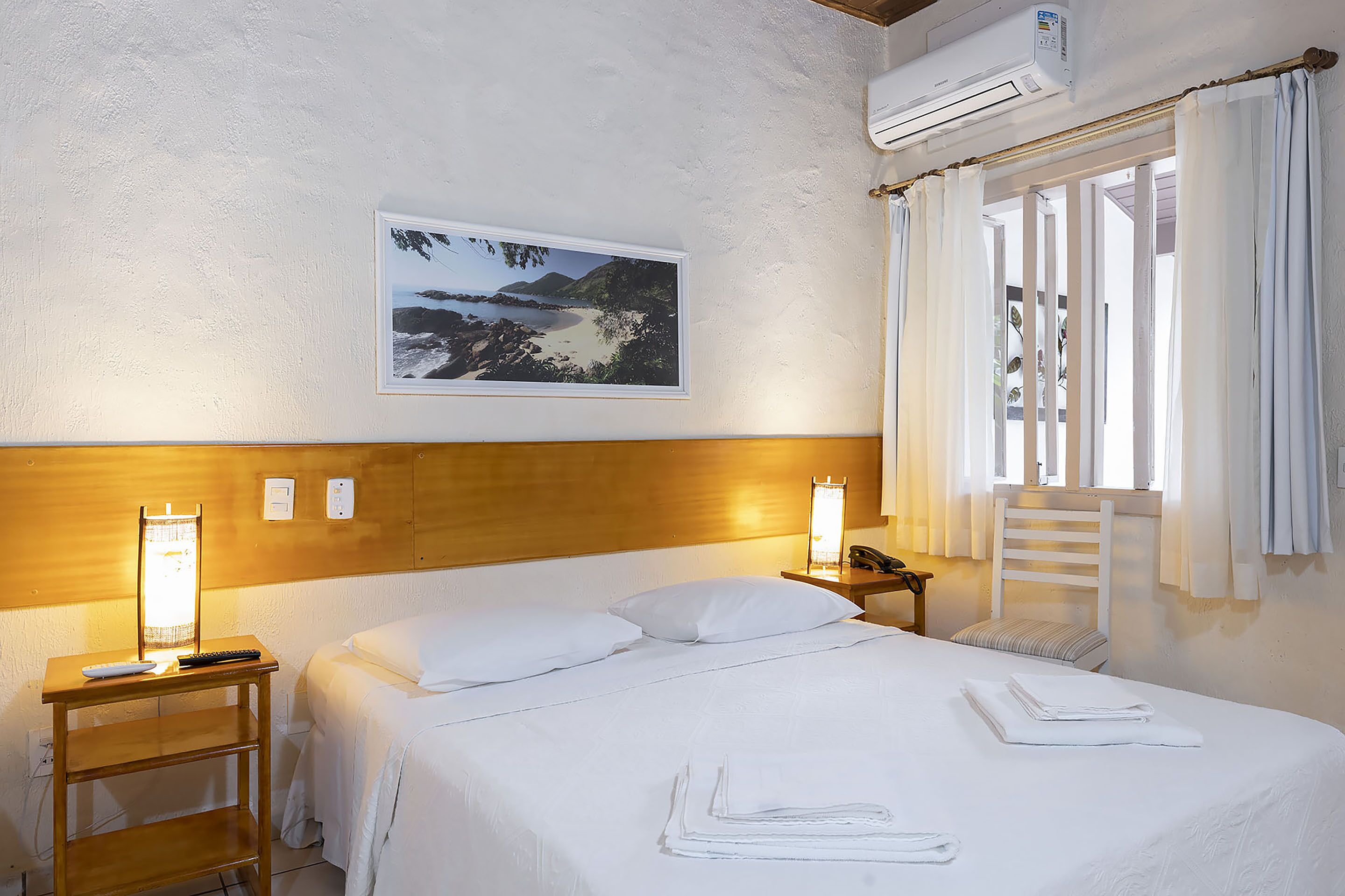 Un lit bien fait, avec du linge de lit blanc, une table de chevet avec une lampe, une commode, un climatiseur fixé au mur et un tableau représentant un paysage.