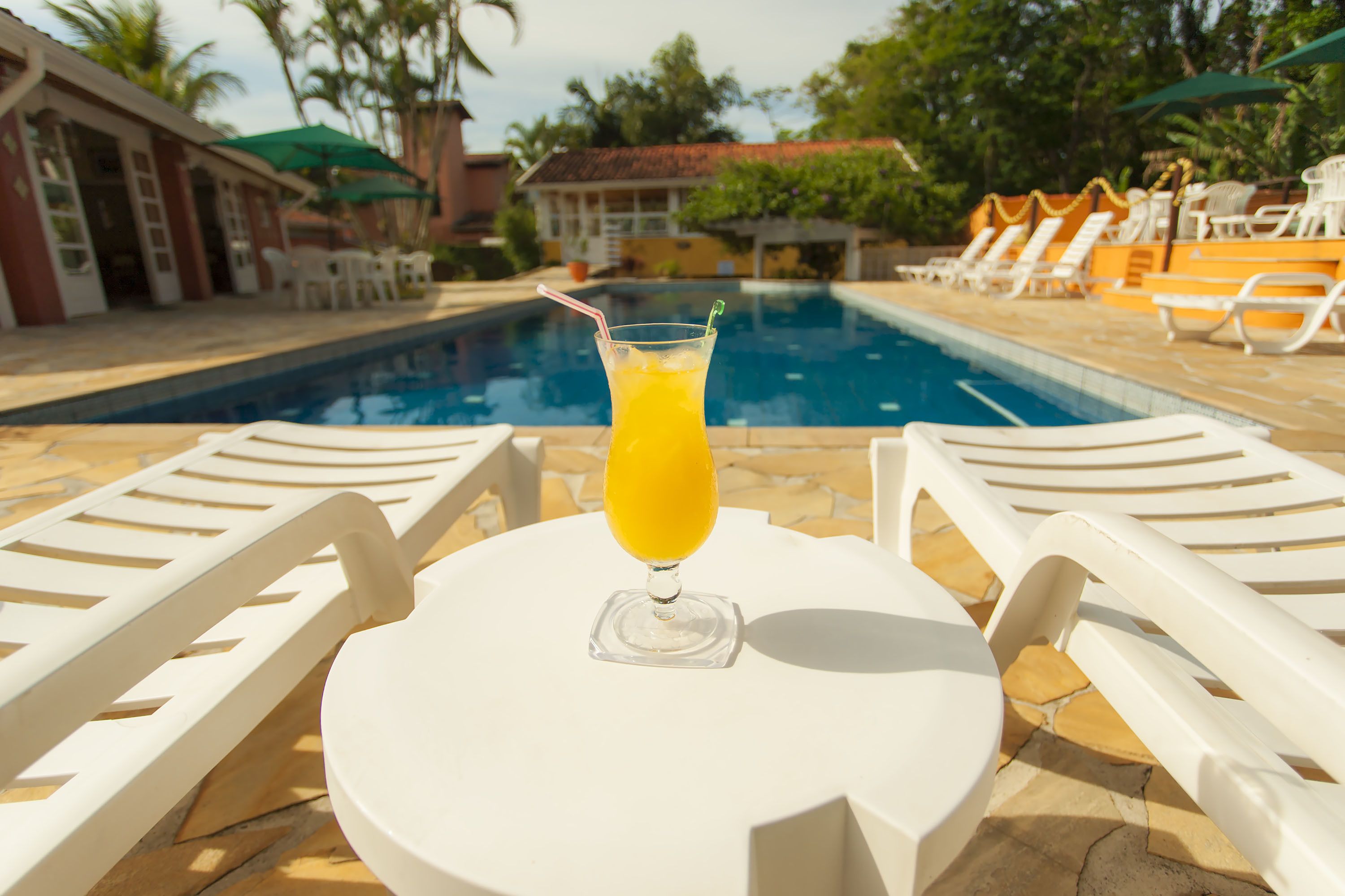 Escena junto a la piscina con una mesa y sillas, un vaso de jugo de naranja y una piscina al fondo.