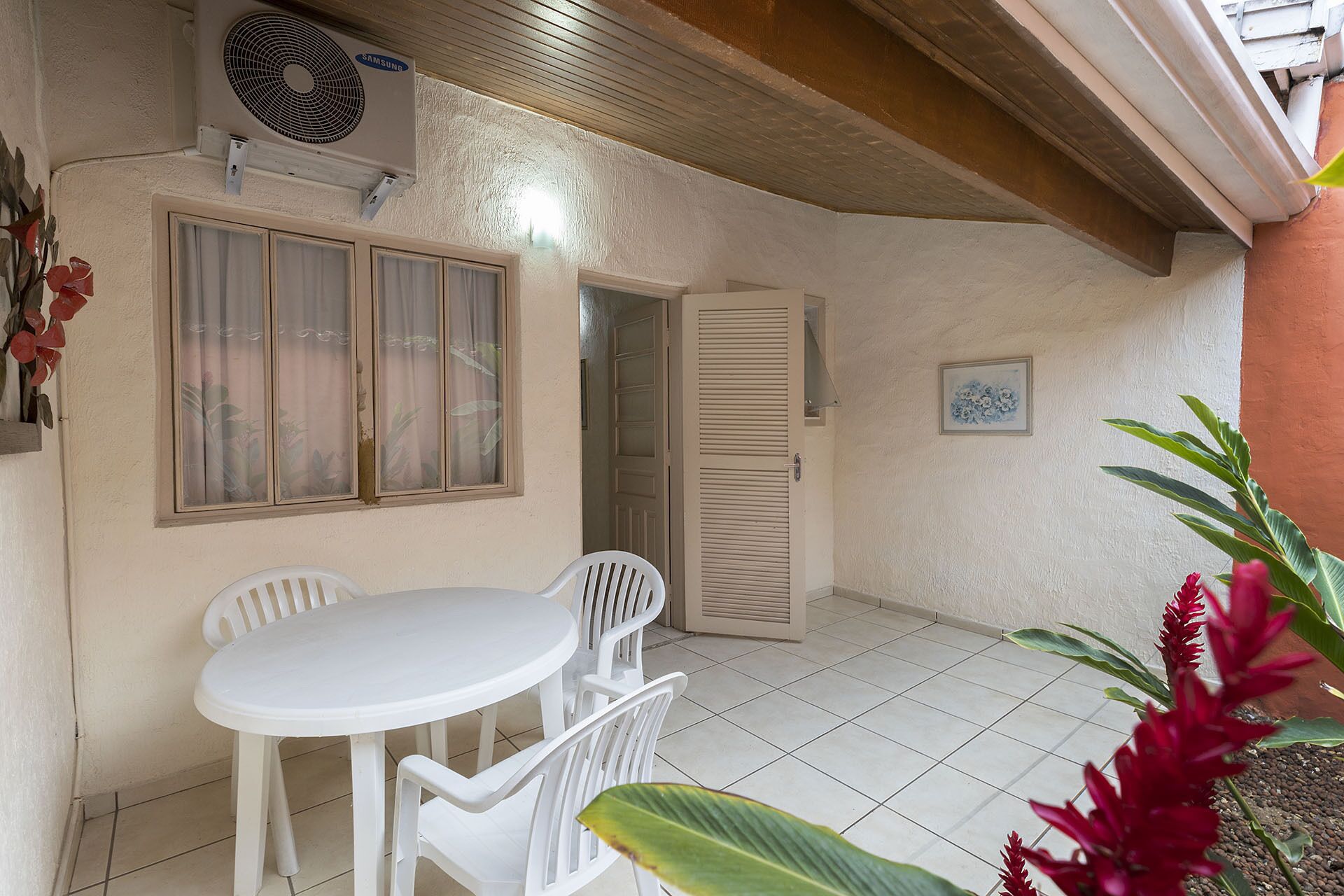 Une petite terrasse avec une table et des chaises blanches, une porte fermée et un ventilateur fixé au mur.