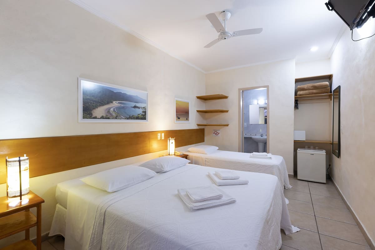 Une chambre d’hôtel avec deux lits, un ventilateur de plafond, un petit réfrigérateur et une salle de bain visible à travers une porte ouverte.