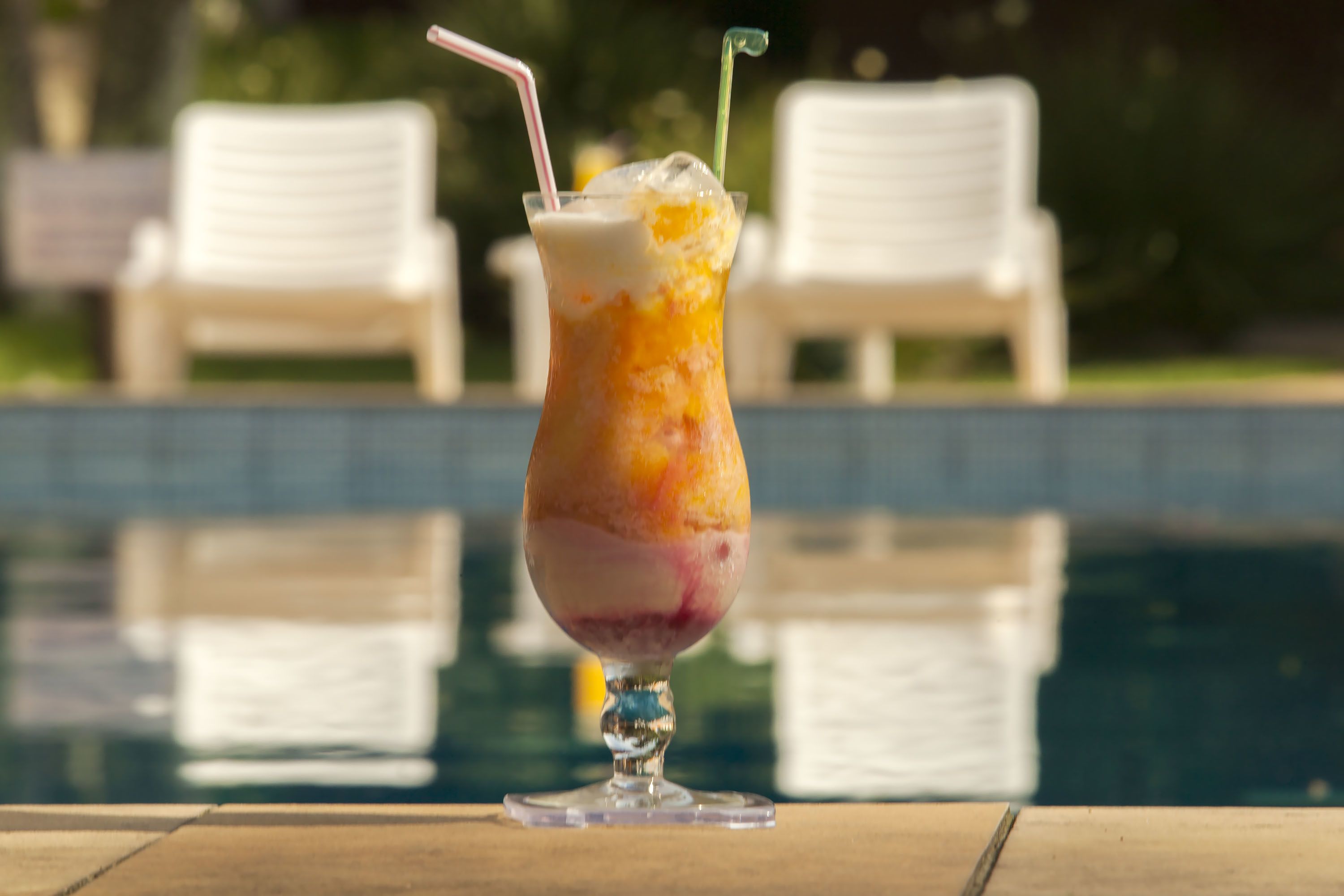 A cocktail with a straw, placed on a table near a poolside with white lounge chairs in the background.