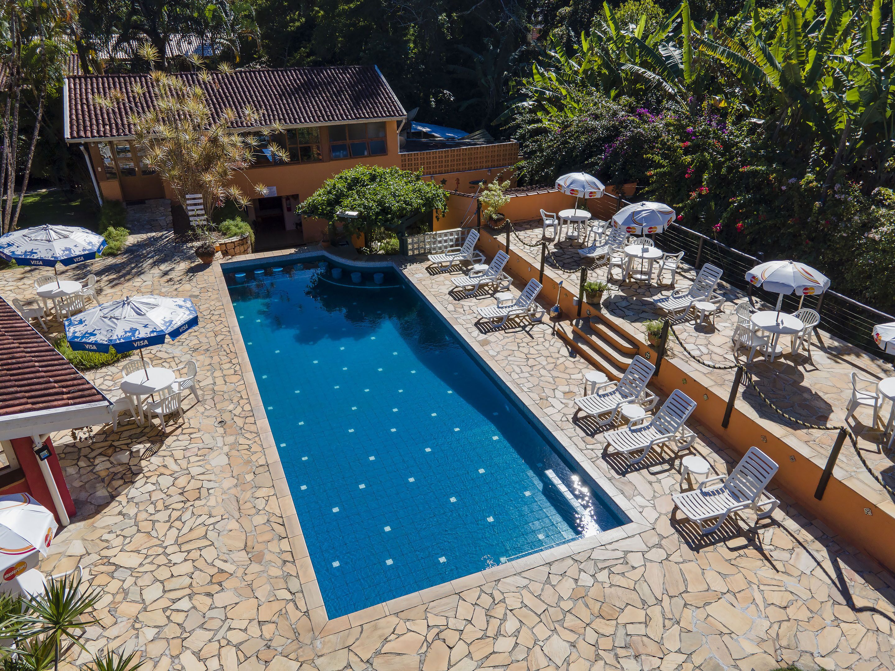 Uma piscina com espreguiçadeiras e guarda-sóis, rodeada por um terraço de pedra e vegetação exuberante.