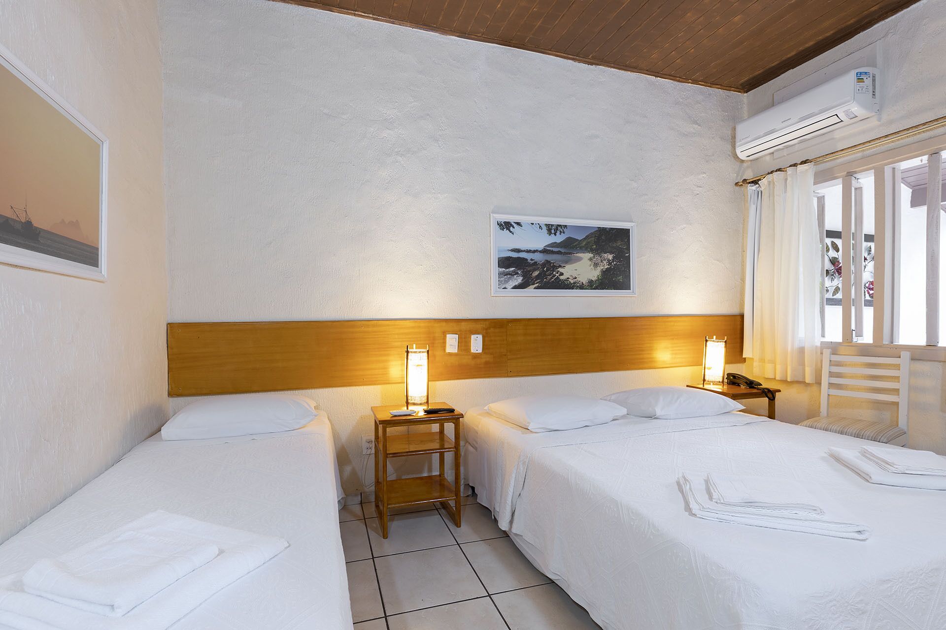 A hotel room with two single beds, a wooden headboard, a nightstand with a lamp, a framed picture on the wall, and an air conditioning unit.