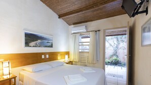 Un lit bien fait, avec du linge de lit blanc, une table de chevet avec une lampe, un tableau encadré au mur et une vue sur un jardin par une porte ouverte.