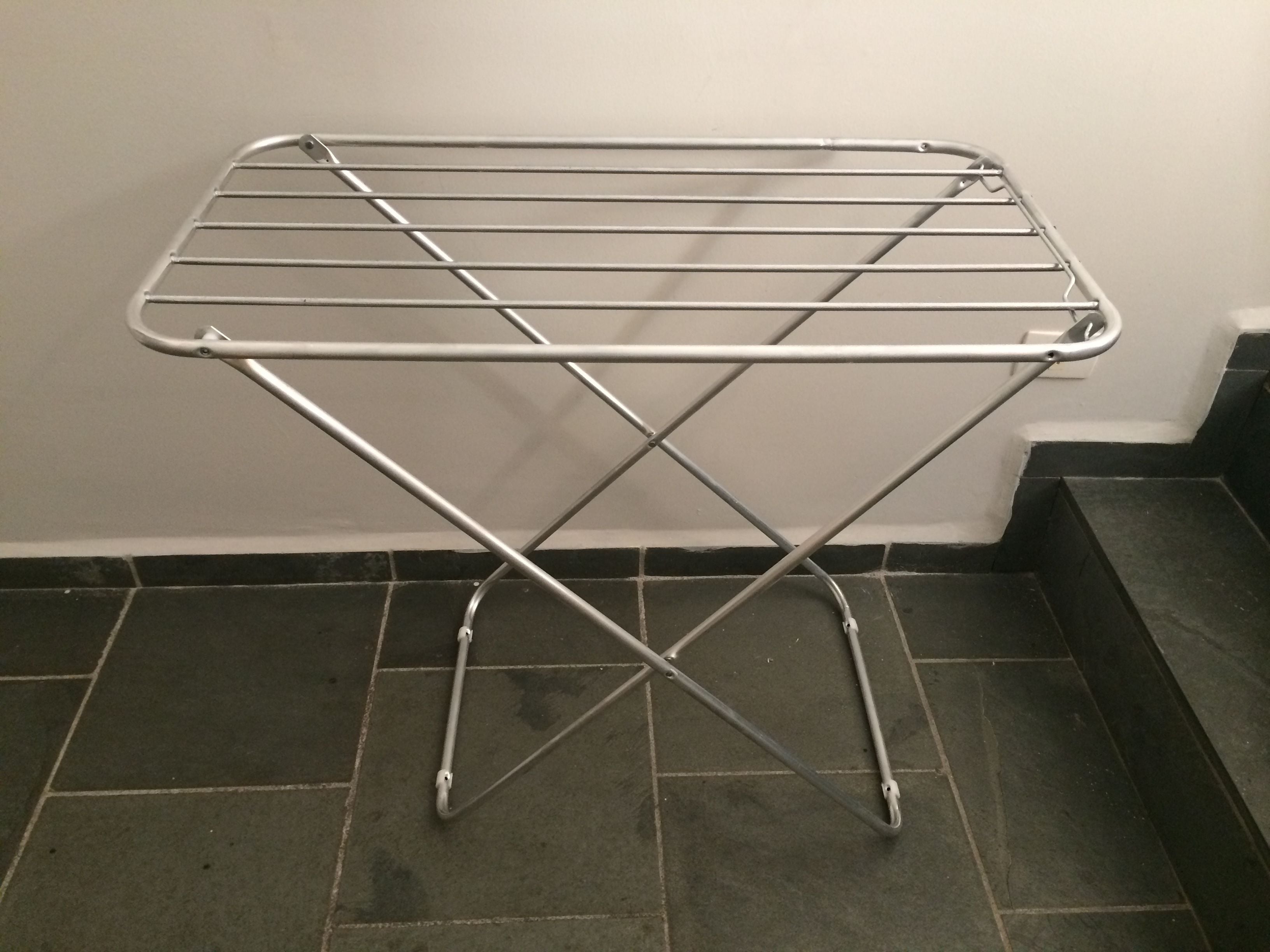 A metal drying rack with a crossbar design, placed on a tiled floor against a plain wall.