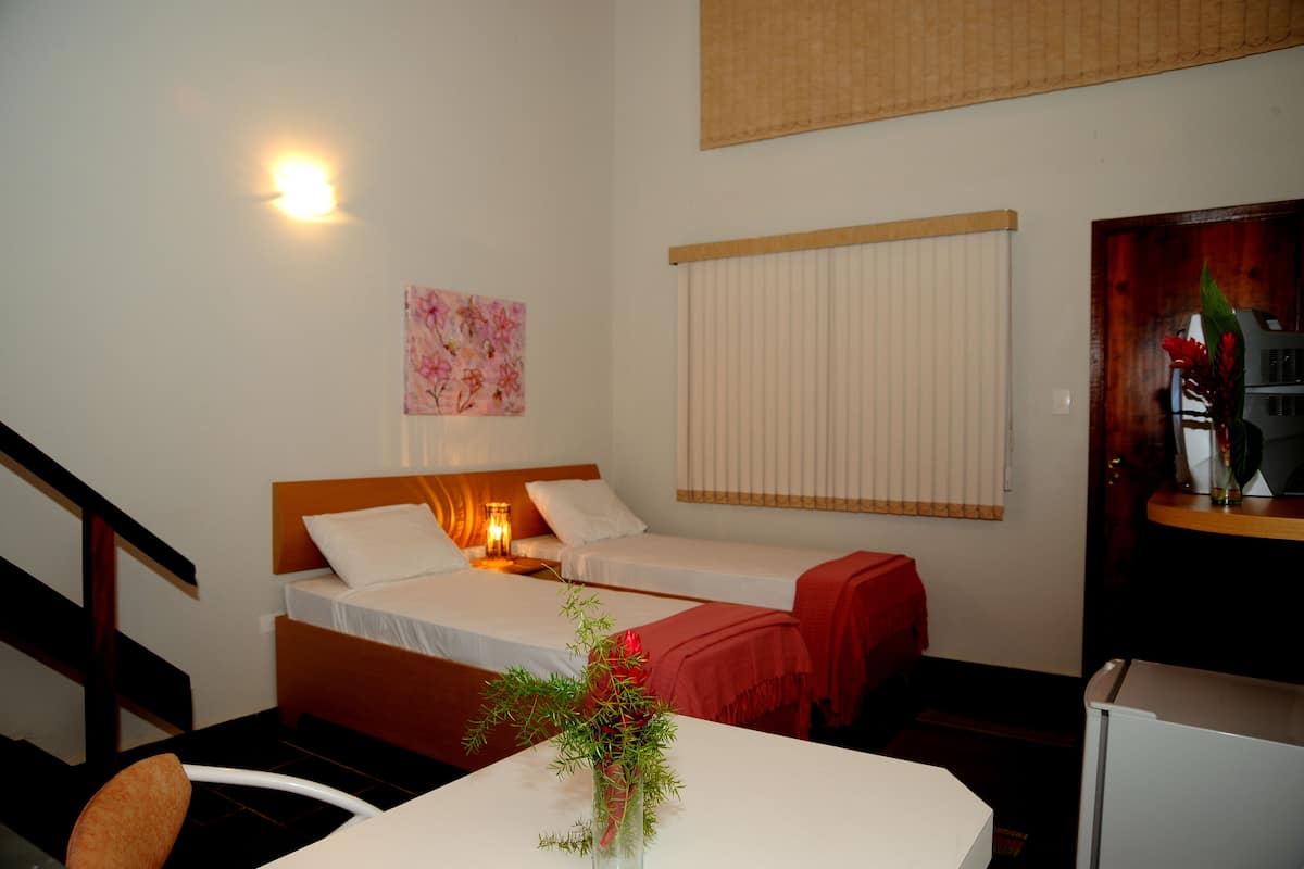 A hotel room with two beds, a nightstand with a lamp, a window with blinds, a painting on the wall, and a small table with a vase of flowers.