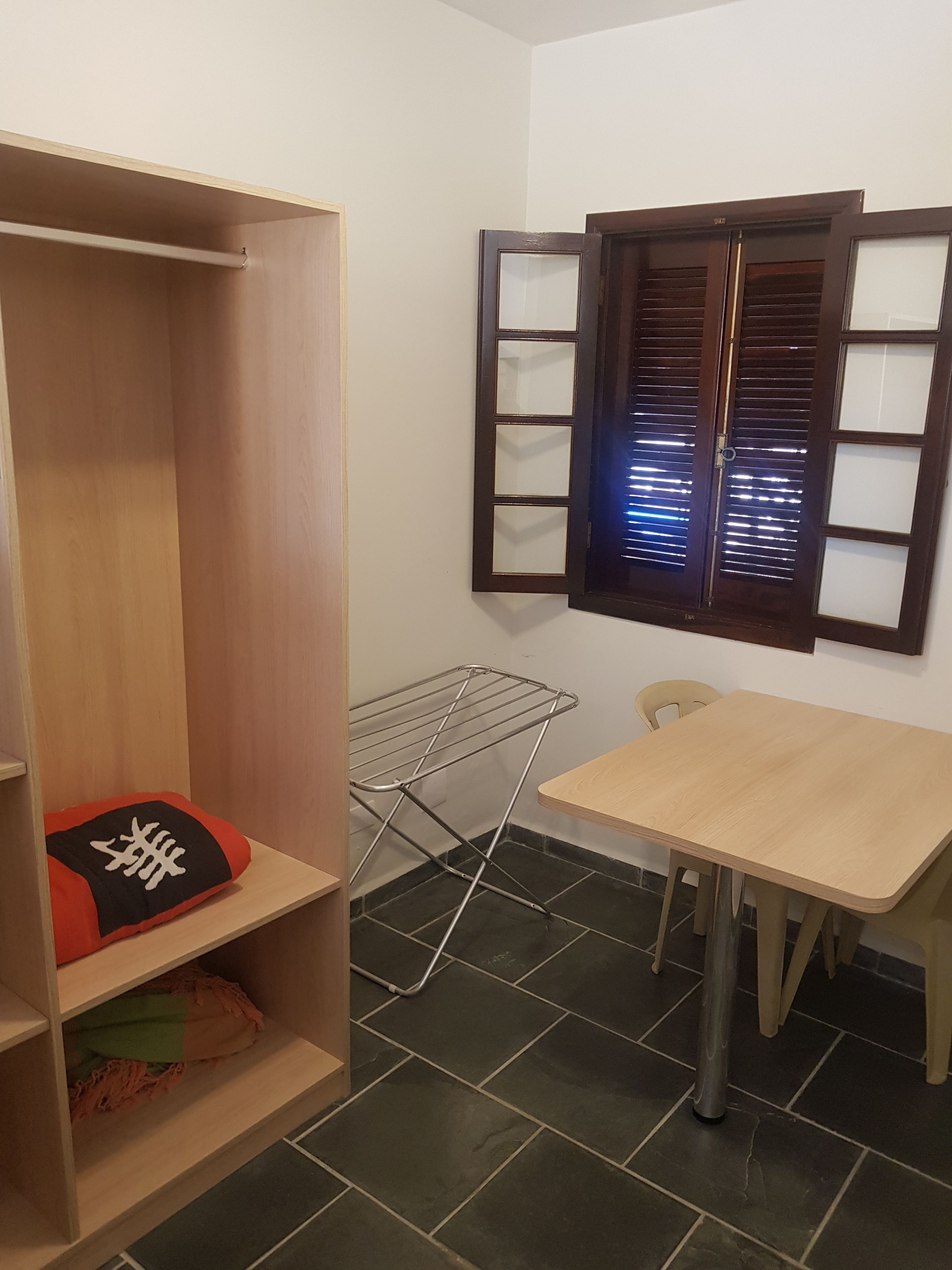 A room with a wooden wardrobe, a small table, a chair, a drying rack, and a window with blinds.