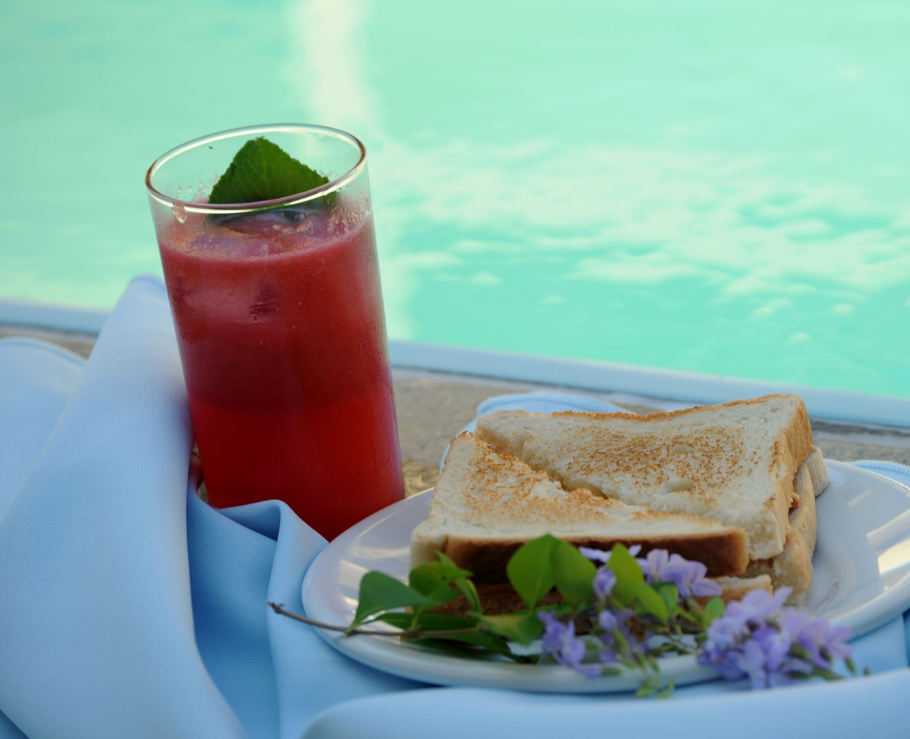 A glass of red beverage with a mint leaf, a sandwich on a plate, and a pool in the background.