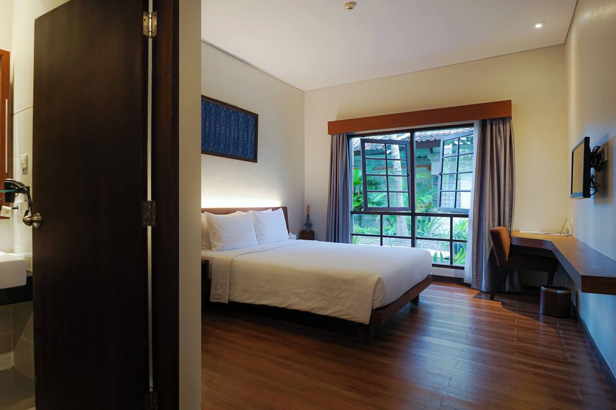 Une chambre d’hôtel avec un grand lit, un bureau, une fenêtre donnant sur un espace verdoyant, une salle de bain avec un lavabo et un miroir.