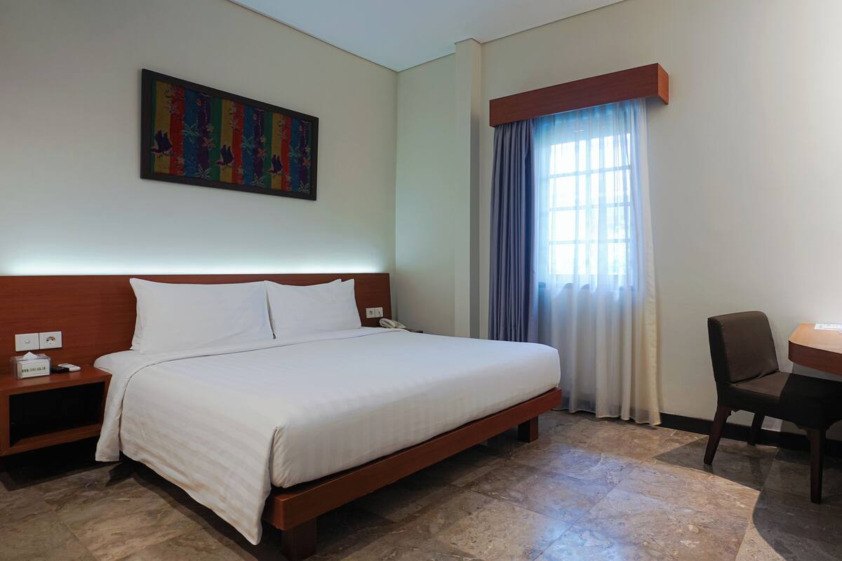 Une chambre d’hôtel avec un lit, un bureau, une chaise, un tableau au mur et une fenêtre avec des rideaux.