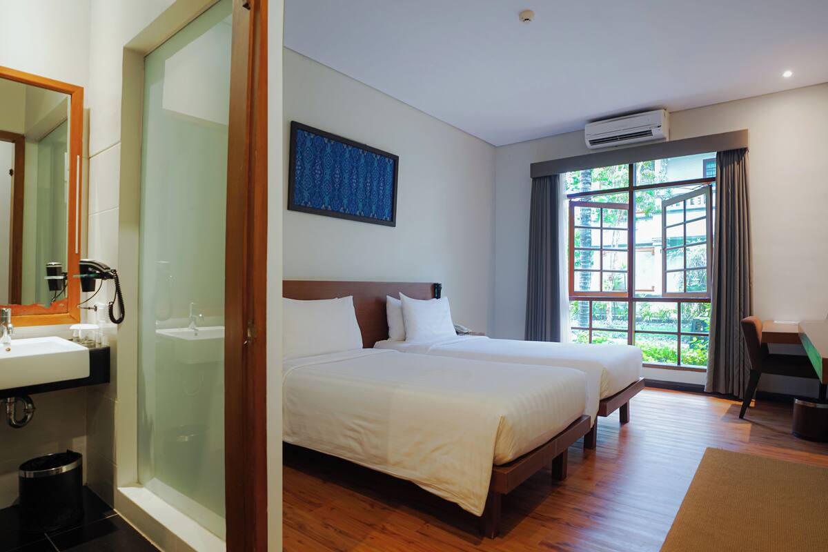 Une chambre d’hôtel avec un grand lit, une table de chevet, un miroir, une fenêtre donnant sur un espace verdoyant et une salle de bain avec un lavabo et un miroir.