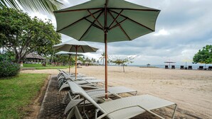 A beachside lounge area with wicker lounge chairs and large umbrellas, a sandy beach, and palm trees in the background. - Grand Whiz Hotel Nusa Dua (Nusa Dua)