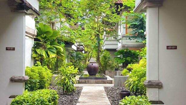 A narrow pathway flanked by lush greenery and a large decorative pot, leading to a building with a traditional architectural style. - Grand Whiz Hotel Nusa Dua (Nusa Dua)