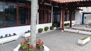 A building with a tiled roof, large windows, and a paved courtyard with planters and a tree. - Hotel Piçarras (Picarras)
