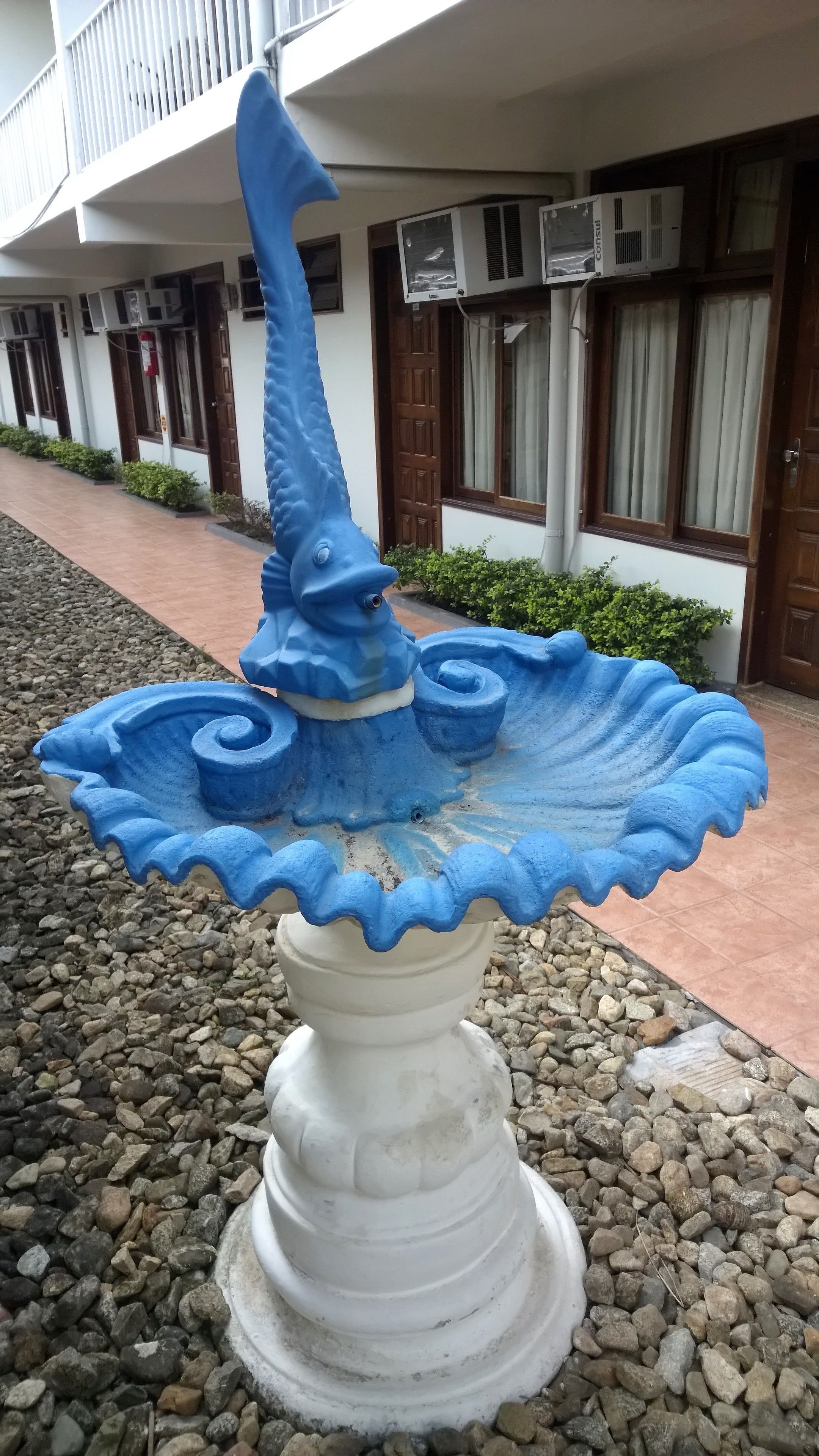 Una fuente decorativa blanca y azul con diseño en espiral, ubicada frente a un edificio con múltiples ventanas y unidades de aire acondicionado.