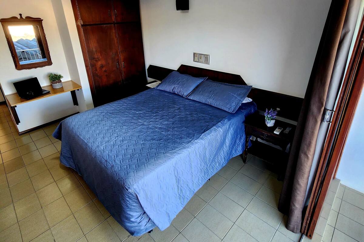 A bedroom with a large bed, a wooden wardrobe, a small desk with a laptop, and a mirror on the wall.