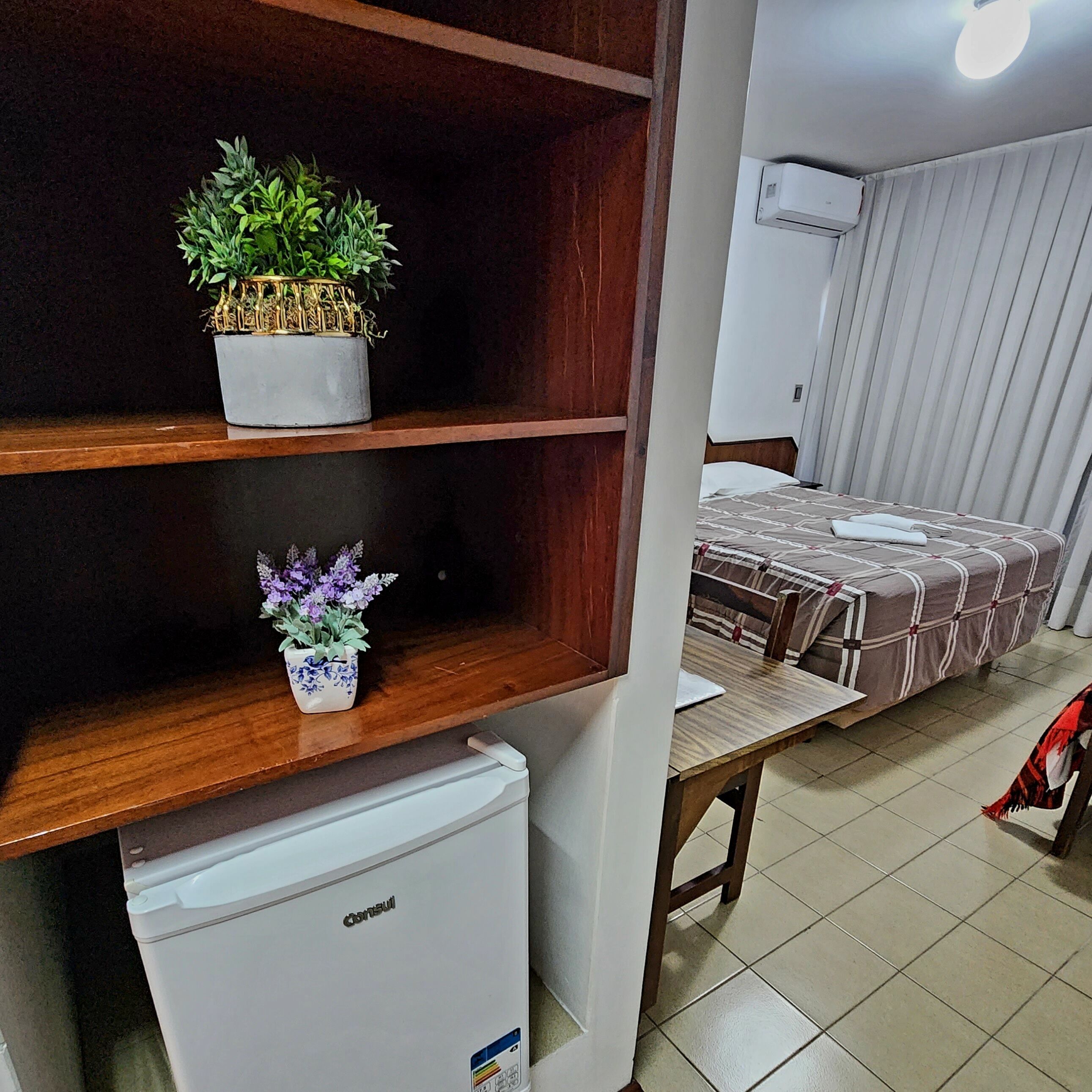Una habitación compacta con una cama, un refrigerador pequeño y dos estantes con plantas en macetas.