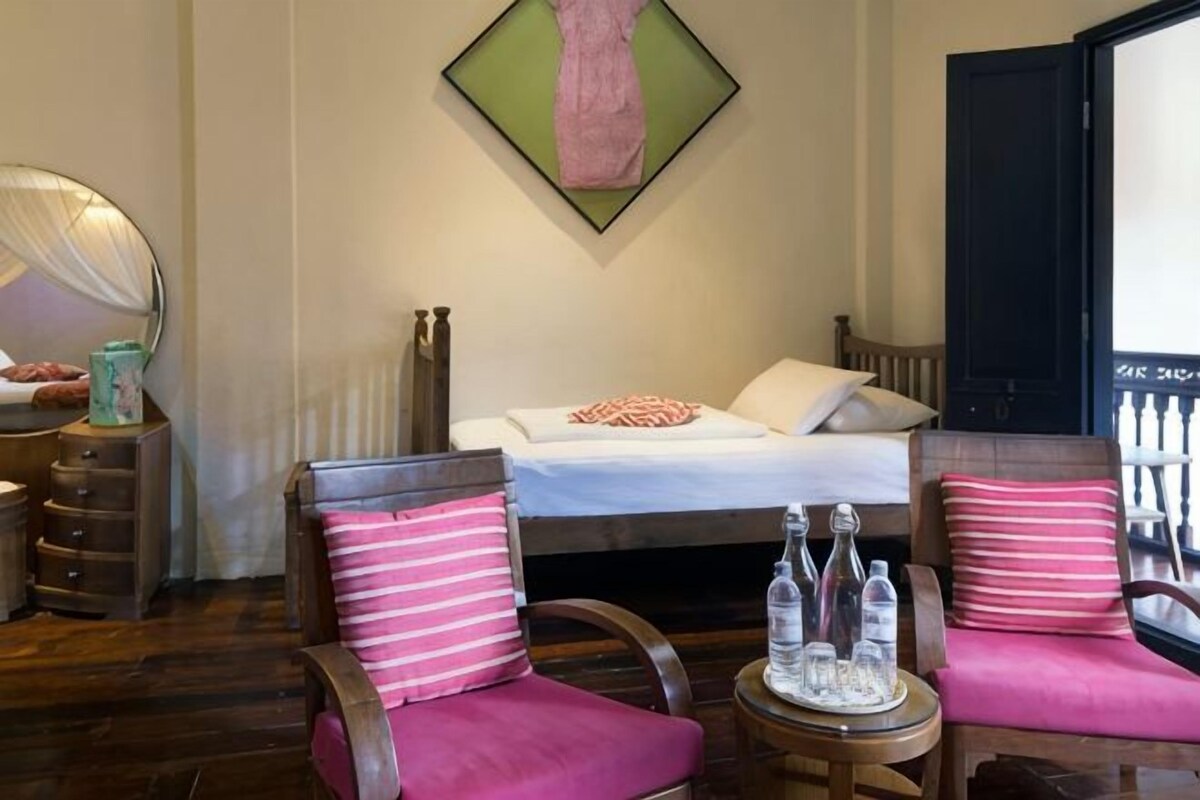 Ein Schlafzimmer mit einem Bett, zwei Stühlen mit rosafarbenen Kissen, einem Nachttisch mit Flaschen, einem Spiegel und einem Wandbild.