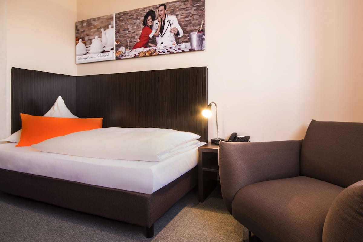 Hotellihuone, jossa on sänky, yöpöytä, lamppu, sohva ja seinä, jossa on kehystetty taulu.