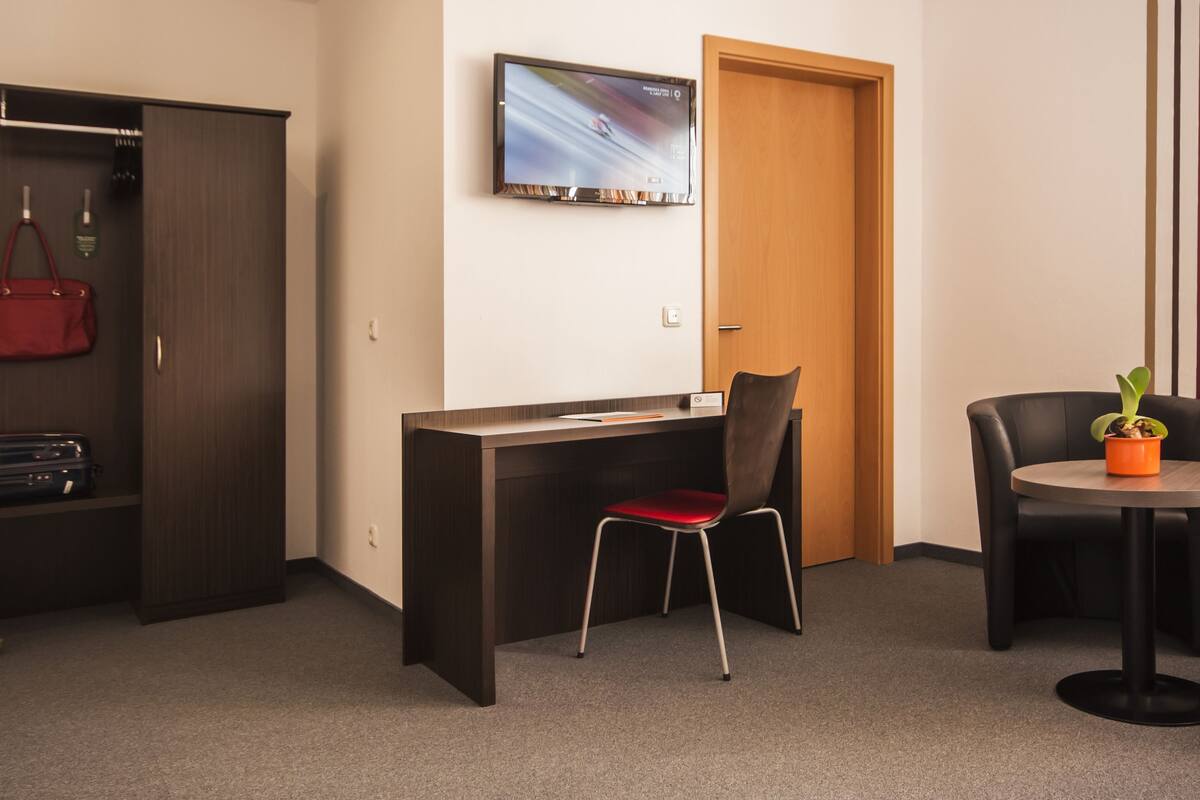 Hotellihuoneessa on työpöytä, tuoli, seinälle asennettu televisio, vaatekaappi ja pieni pöytä, jossa on kasvi.