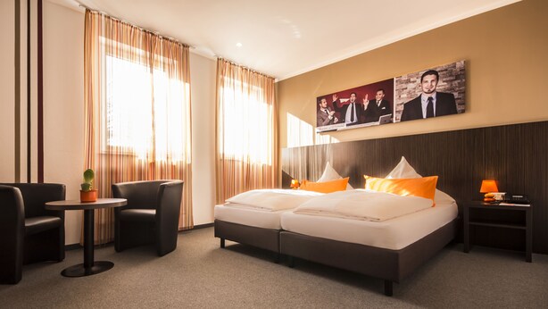 Hotelkamer met een bed, twee stoelen, een klein tafeltje, een nachtkastje en een aan de muur hangend schilderij.