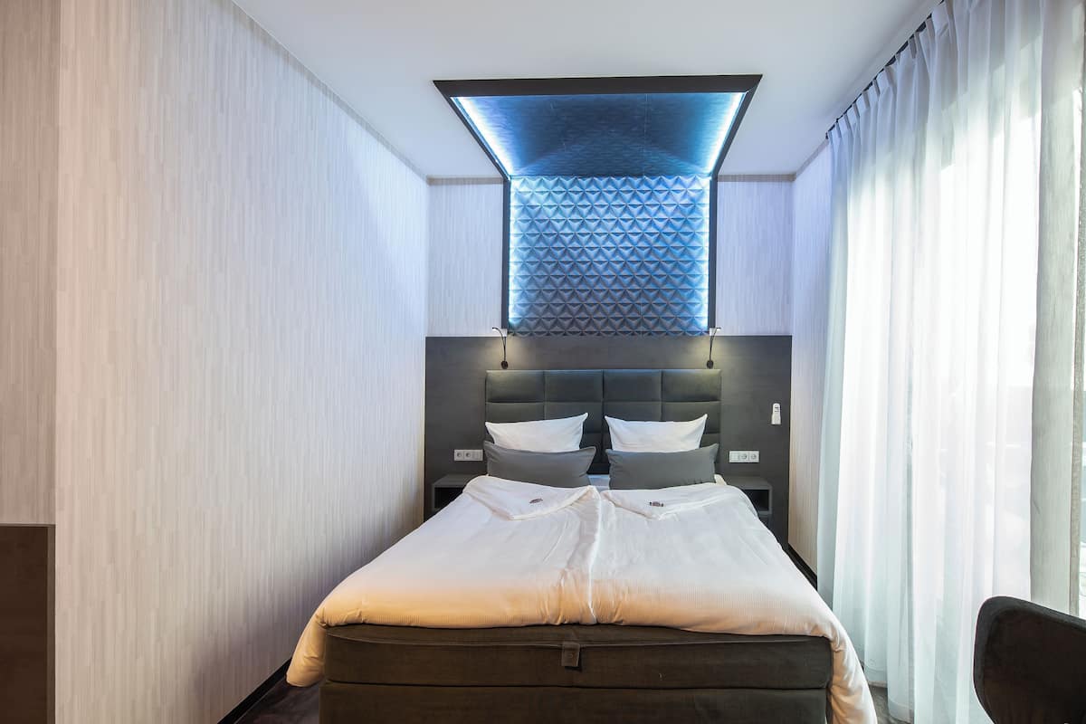 Moderni hotellihuone, jossa on suuri sänky, geometrisesti kuvioitu valaistu pääty ja näkymä ulos ohuiden verhojen läpi.