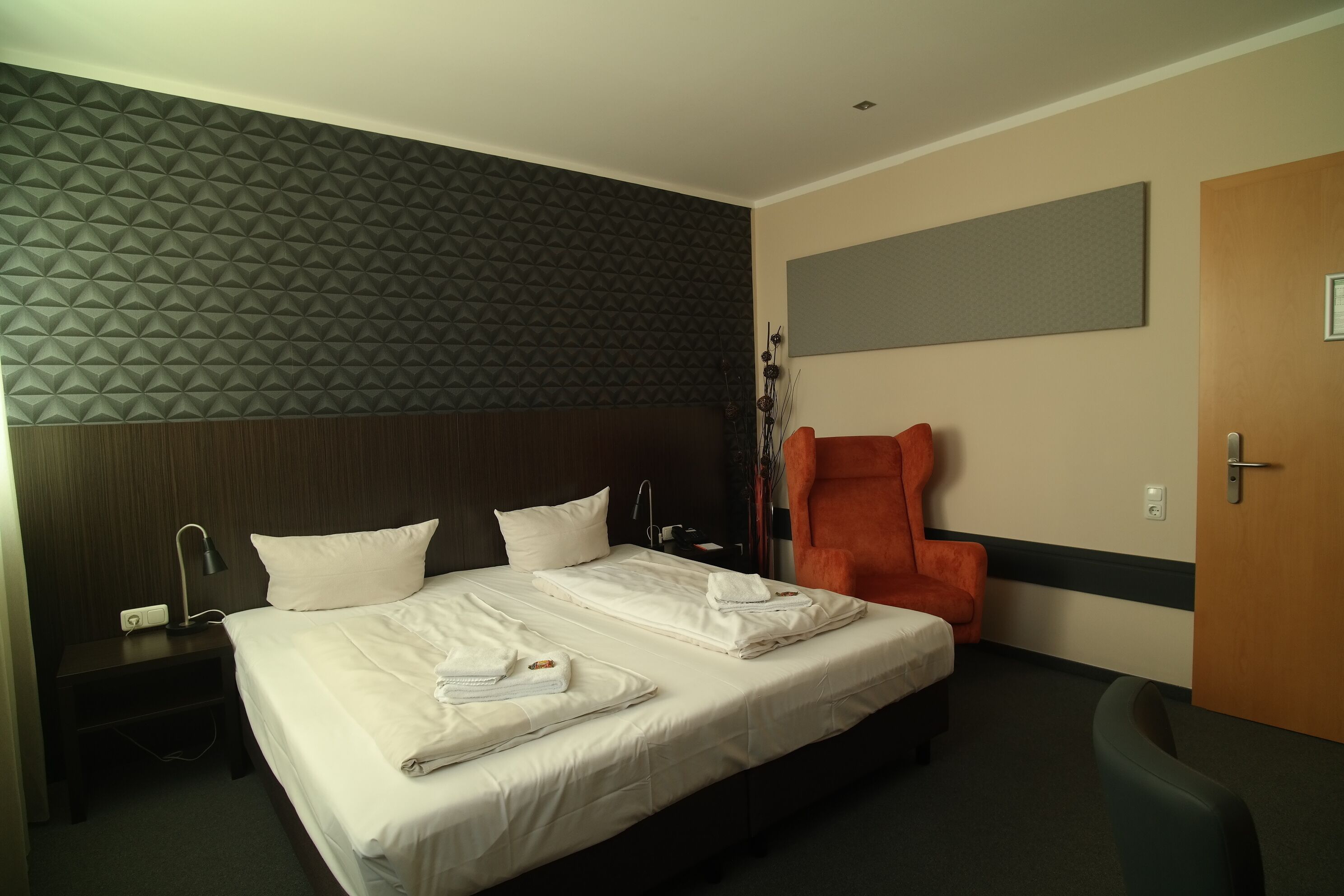 Una camera d'albergo con un letto rifatto, un comodino con una lampada, una sedia e una porta.