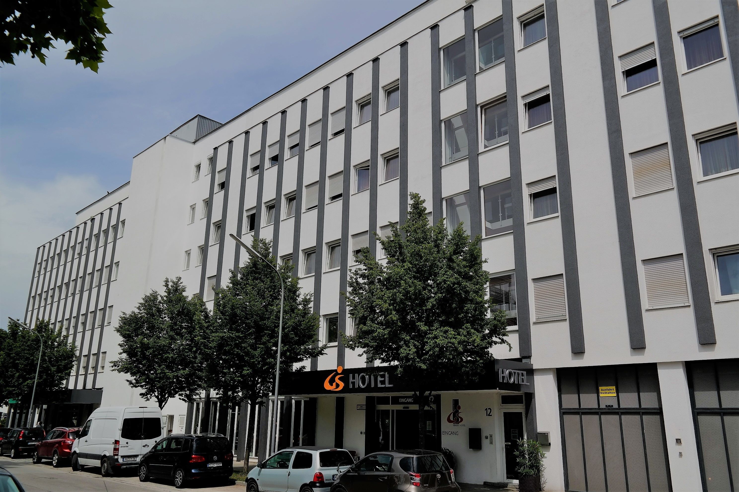 Un moderno edificio de hotel con fachada blanca y numerosas ventanas. Varios automóviles están estacionados en la parte delantera, y se observan algunos árboles.