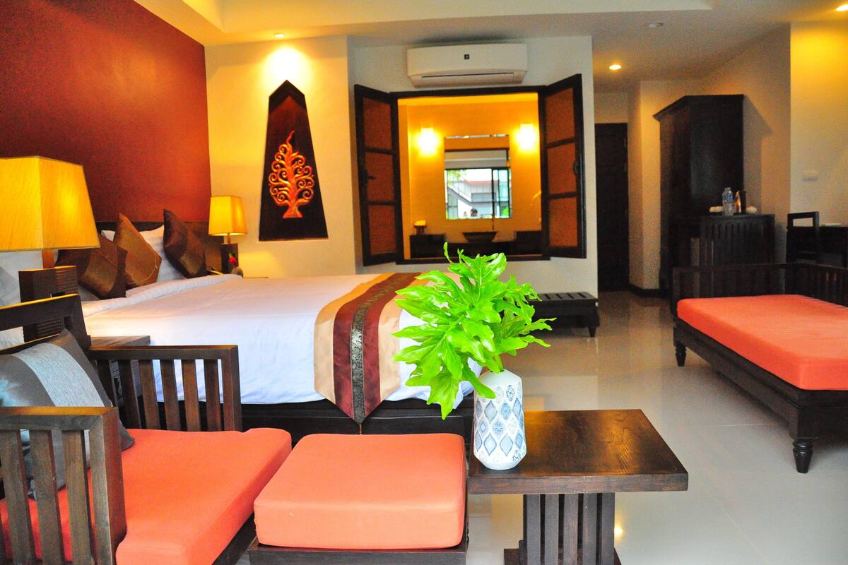 A hotel room with a bed, two orange chairs, a wooden table, and a potted plant.