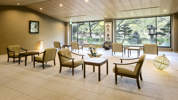 A spacious room with wooden ceiling panels, large windows offering a view of trees and a traditional lantern, and several chairs and tables arranged for seating. - Seizansou (Uji)