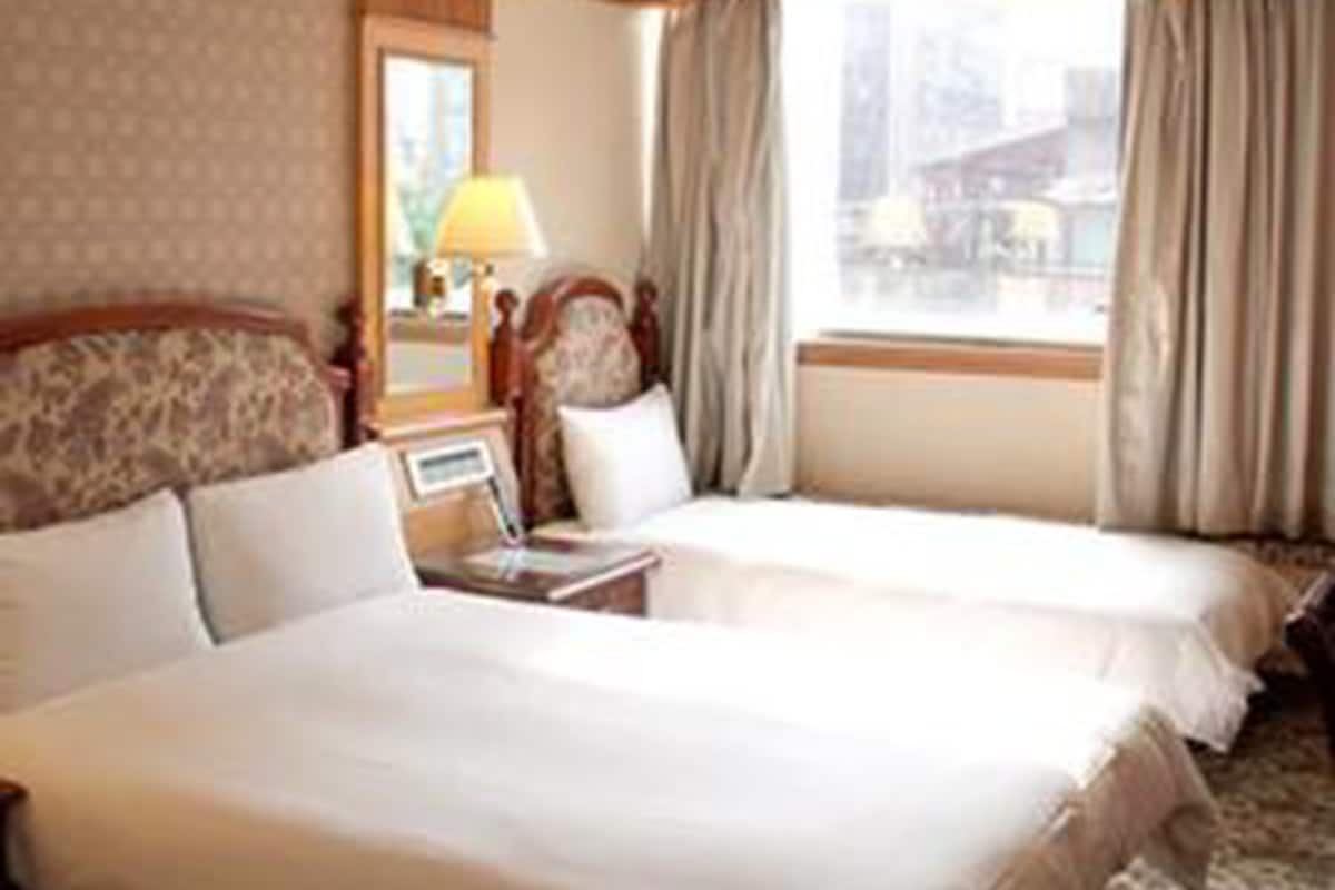 A hotel room with two beds, a bedside table with a lamp, and a window with curtains.
