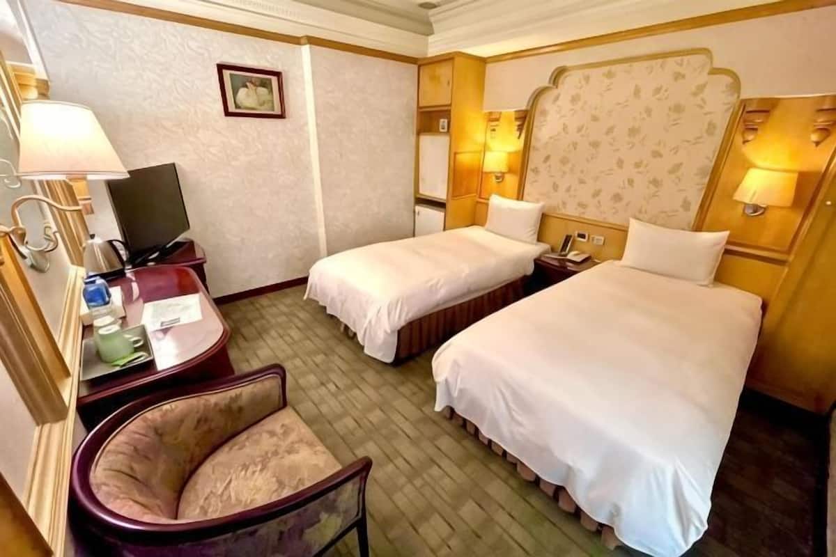 A hotel room with two beds, a desk with a lamp, a chair, and a television.