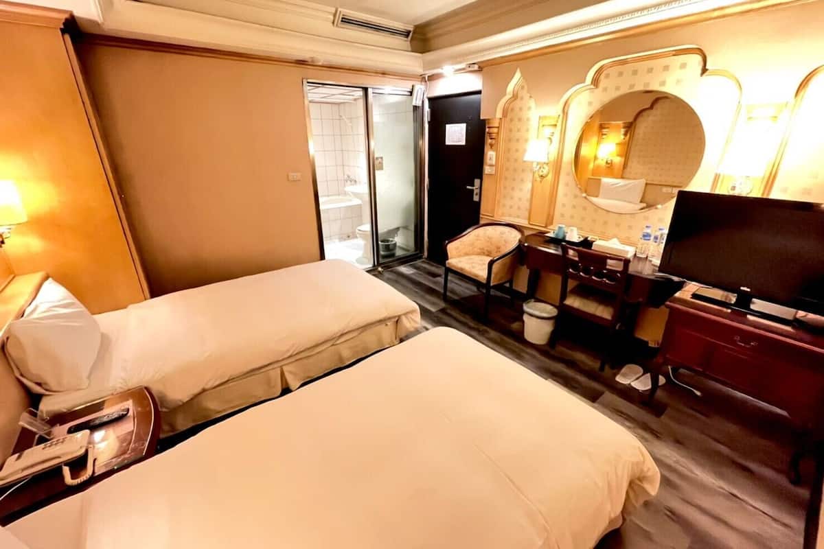 A hotel room with two beds, a desk with a chair, a television, and a bathroom visible through an open door.