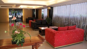Uma sala de estar ampla com um sofá vermelho, poltronas pretas e uma mesa de centro de madeira. Há uma janela grande com cortinas leves e um vaso de planta no parapeito.