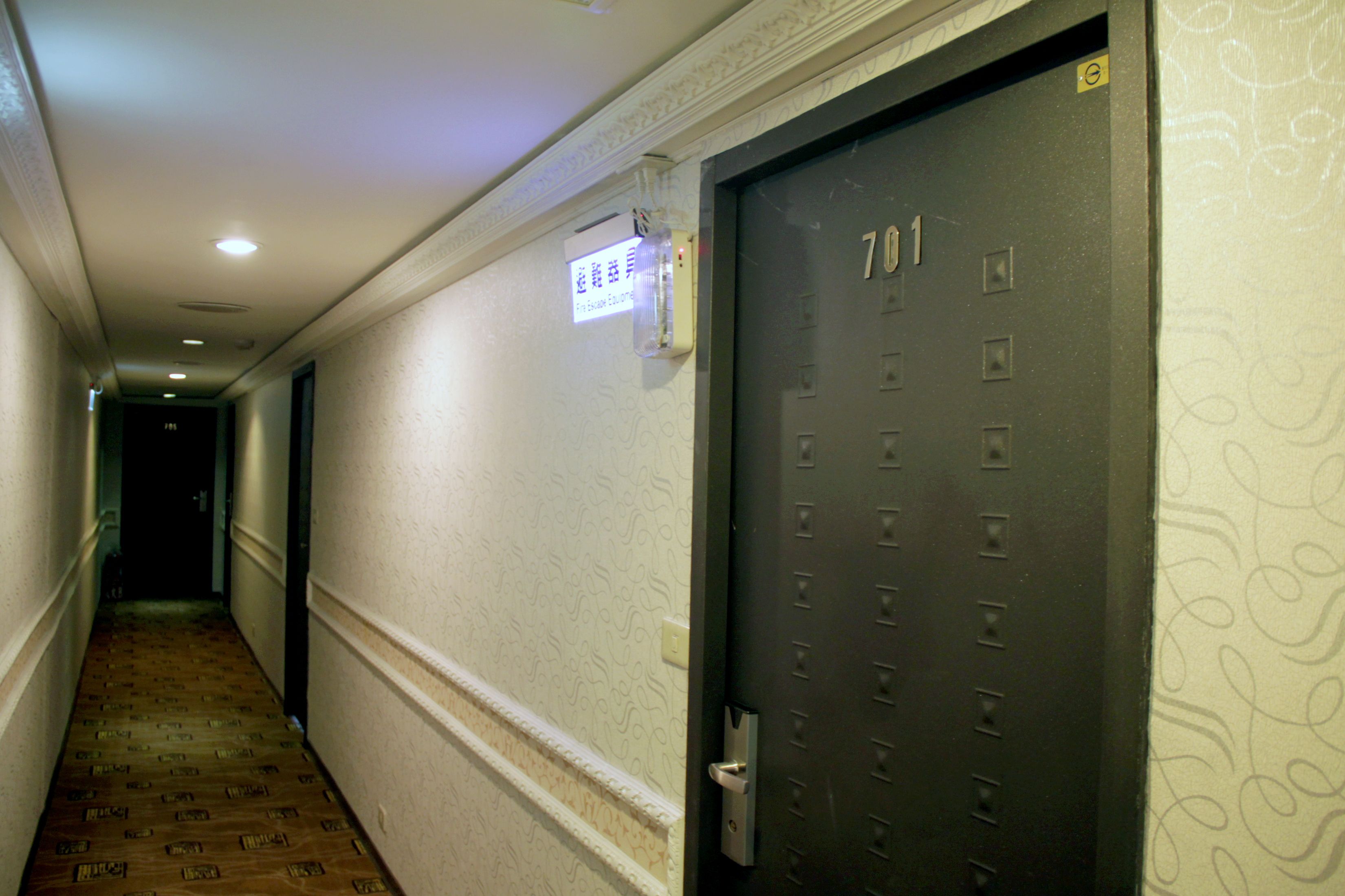 Um corredor com parede estampada, uma porta com a inscrição "701" e um interruptor de luz na parede.