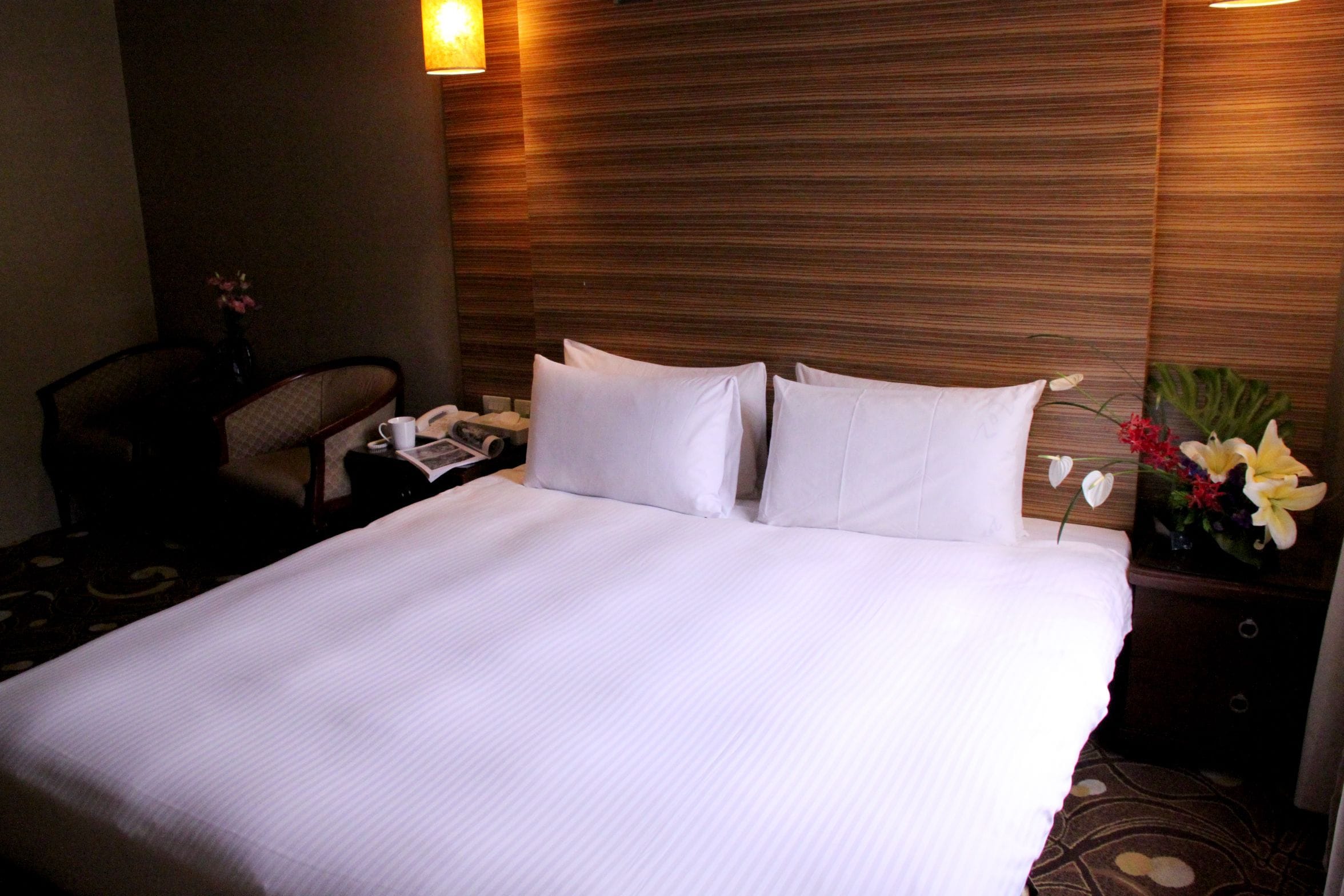 Habitación de hotel con una cama grande, una mesita de noche con lámpara, una silla y una pequeña mesa con un jarrón de flores.
